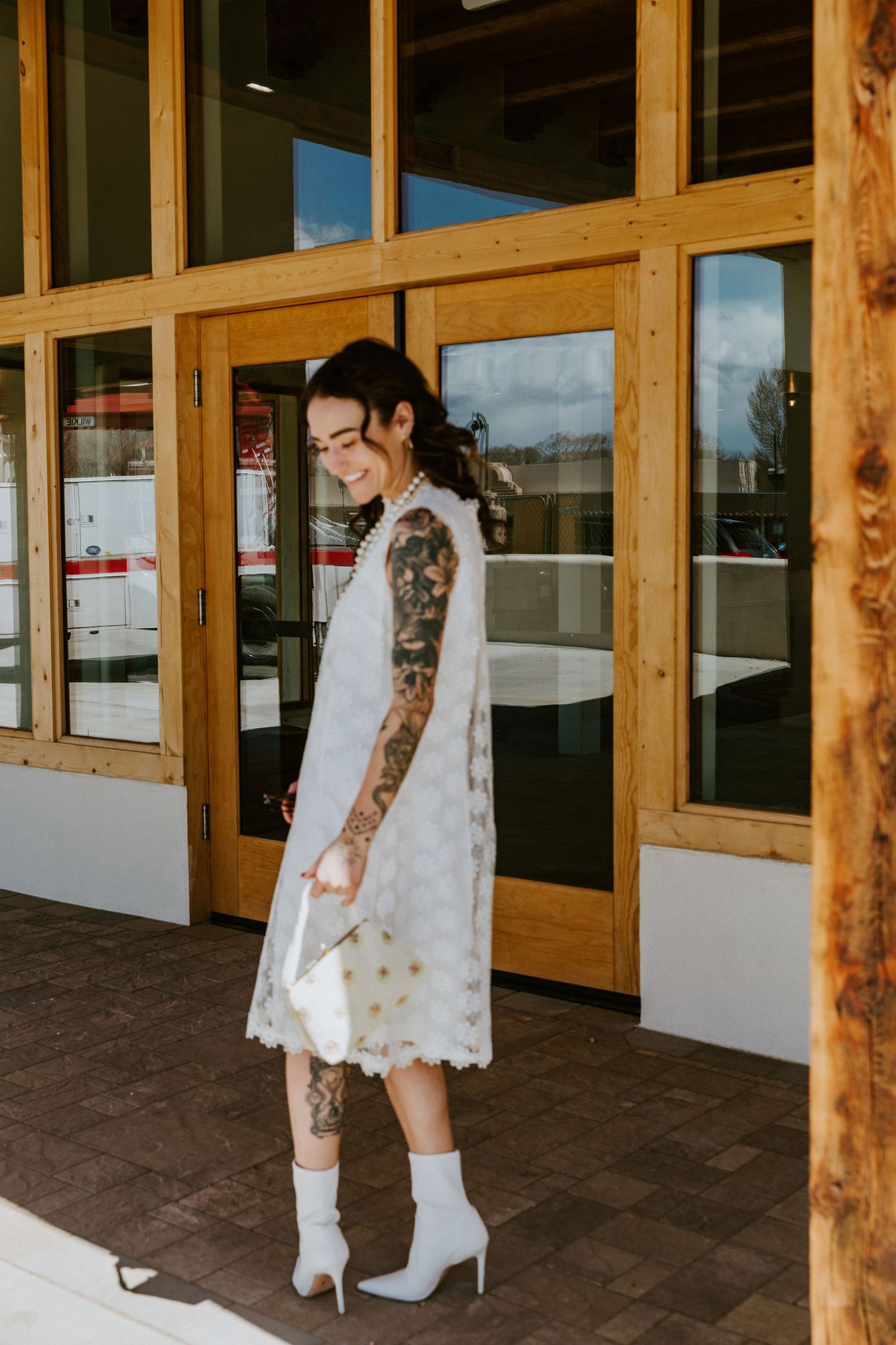 A person in a white polka dot dress and white boots poses near wooden-framed glass doors.