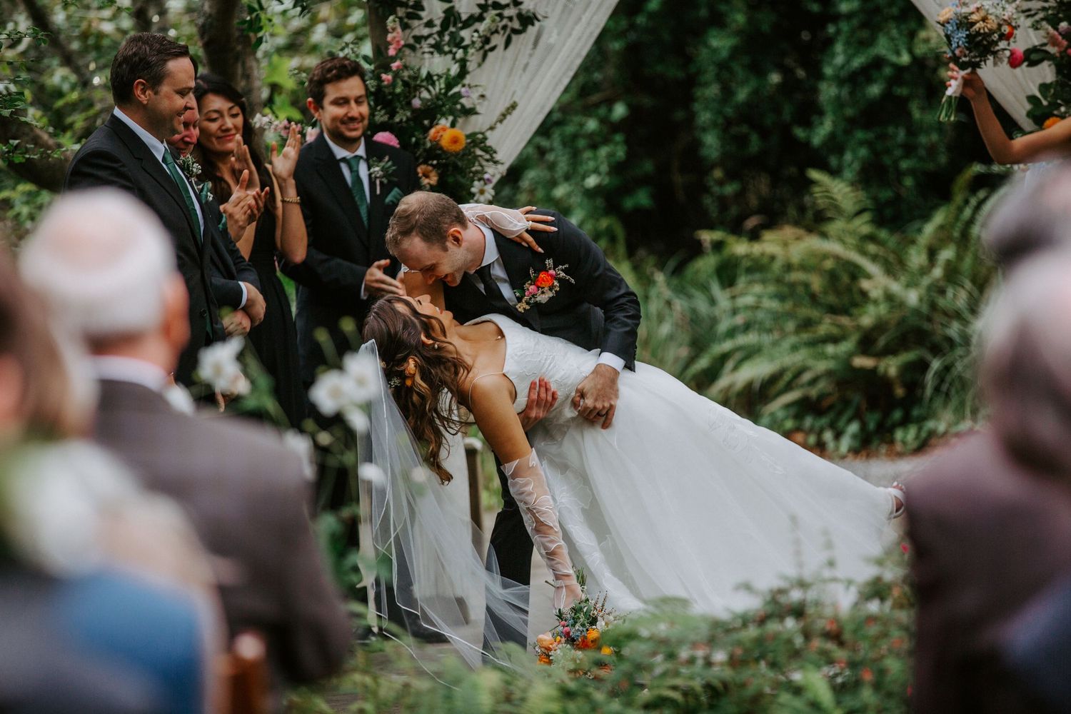 Wedding guests celebrate and cheer during an outdoor garden ceremony under lush green trees.