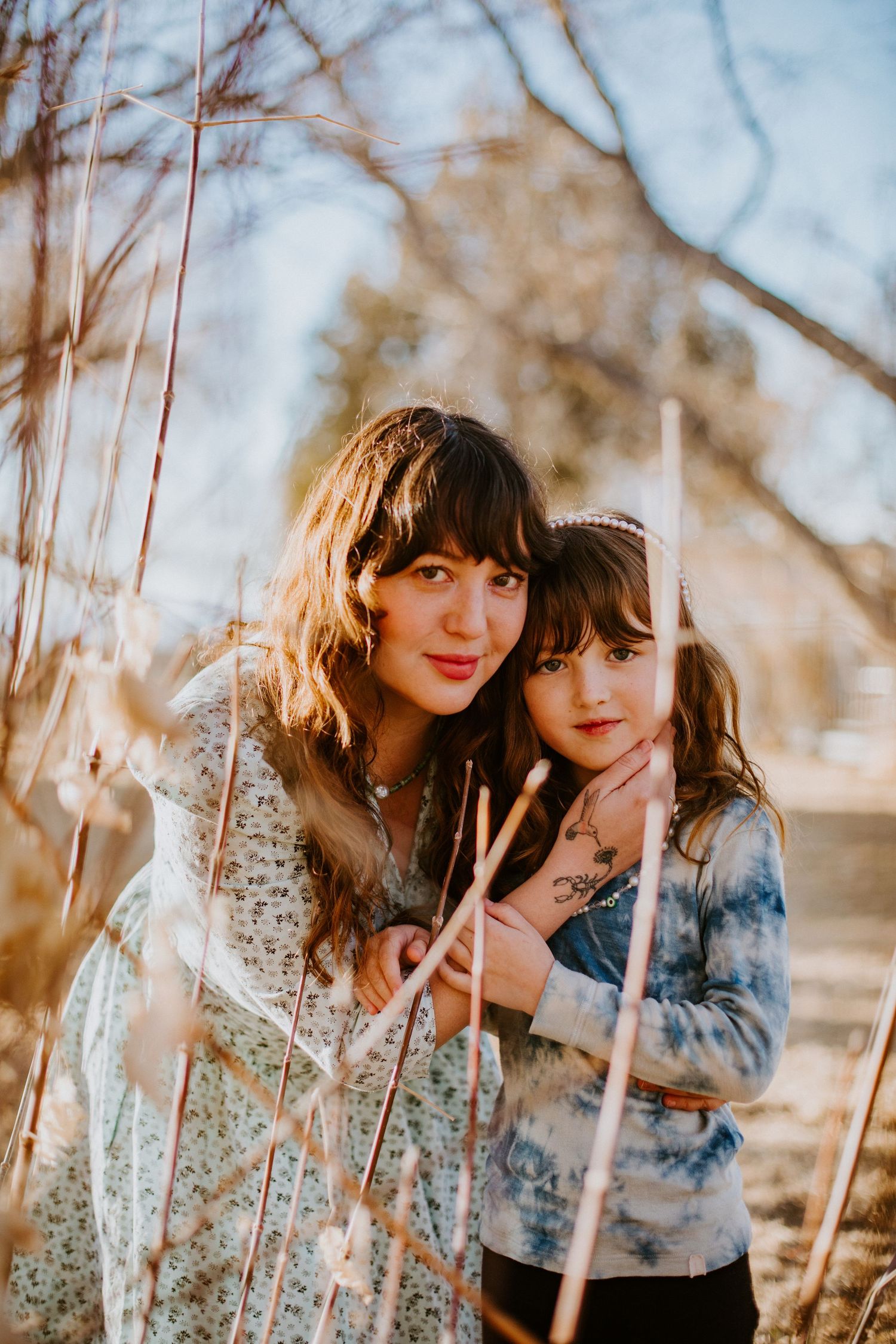 Two people in casual clothing pose together among bare tree branches on a sunny winter day.