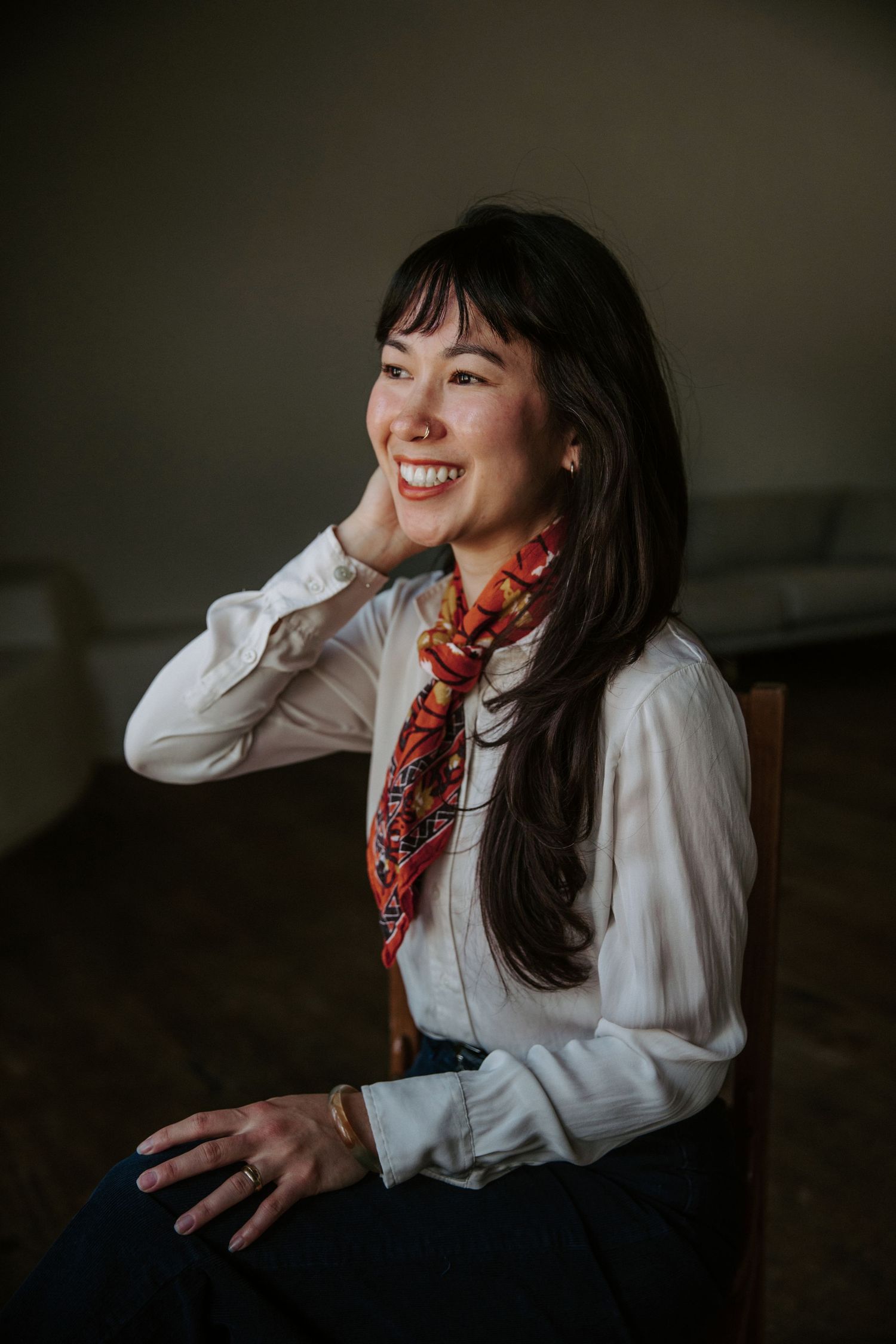Smiling Asian woman in white blouse and colorful scarf, sitting relaxed in chair with hand on cheek.