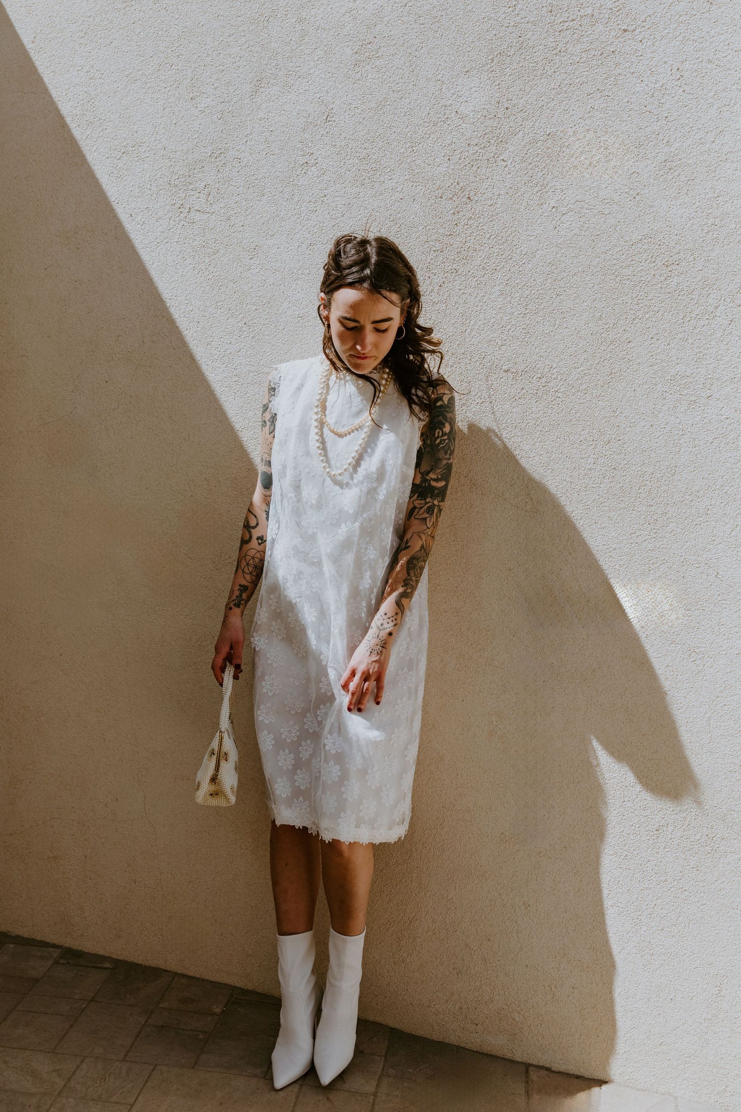 Person wearing a white lace dress stands against a white wall casting dramatic angular shadows.