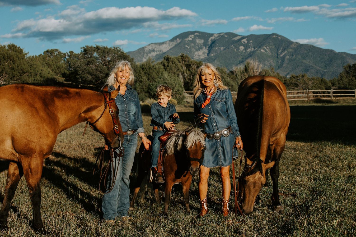 Two people stand with horses in a field with mountains in the background.
