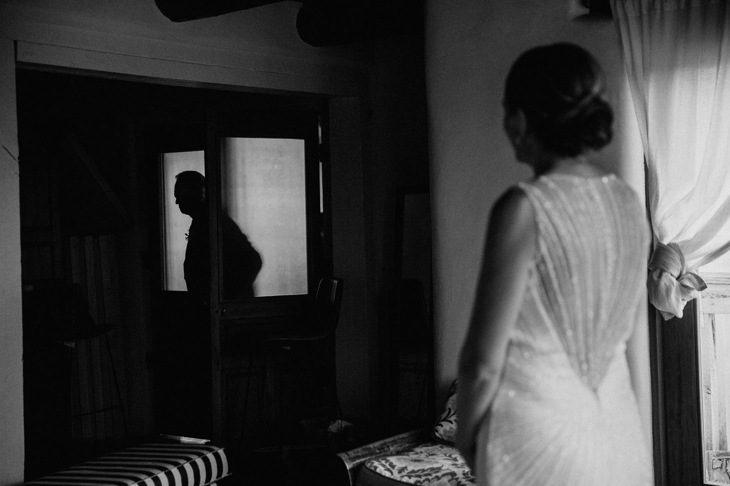 Black and white photo of a bride in a lace dress looking at a silhouette through a doorway.  Shot in Taos, NM by Siouxzen Kang