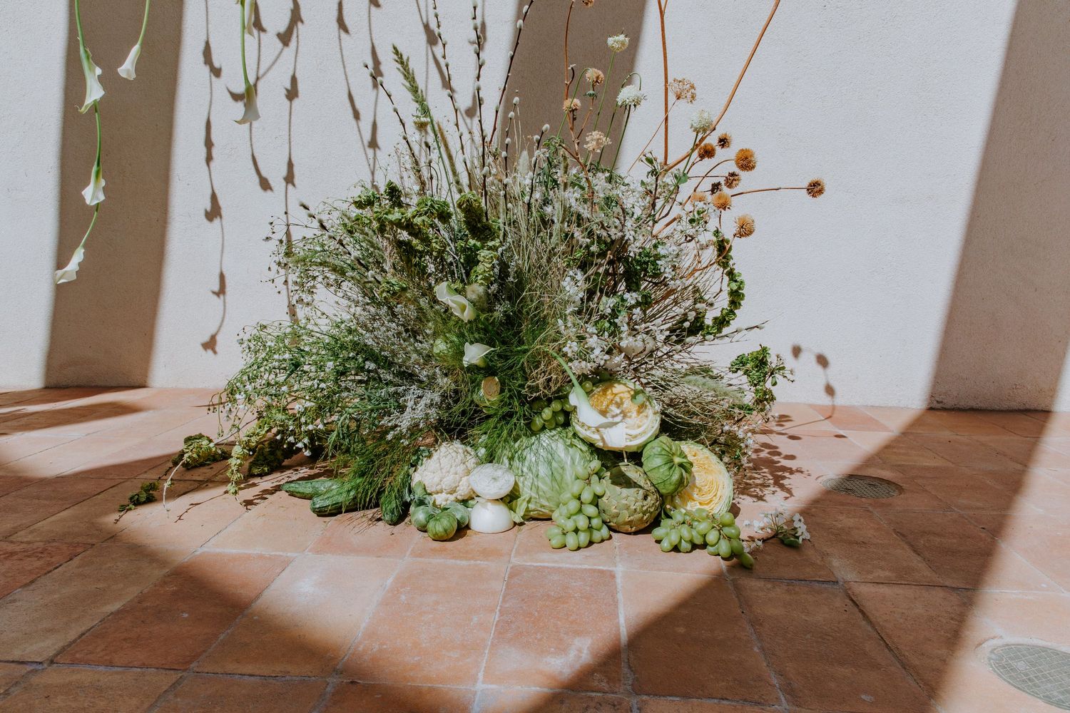 White floral arrangements in glass vases decorate a rustic tiled floor with sunlight casting shadows on the wall.