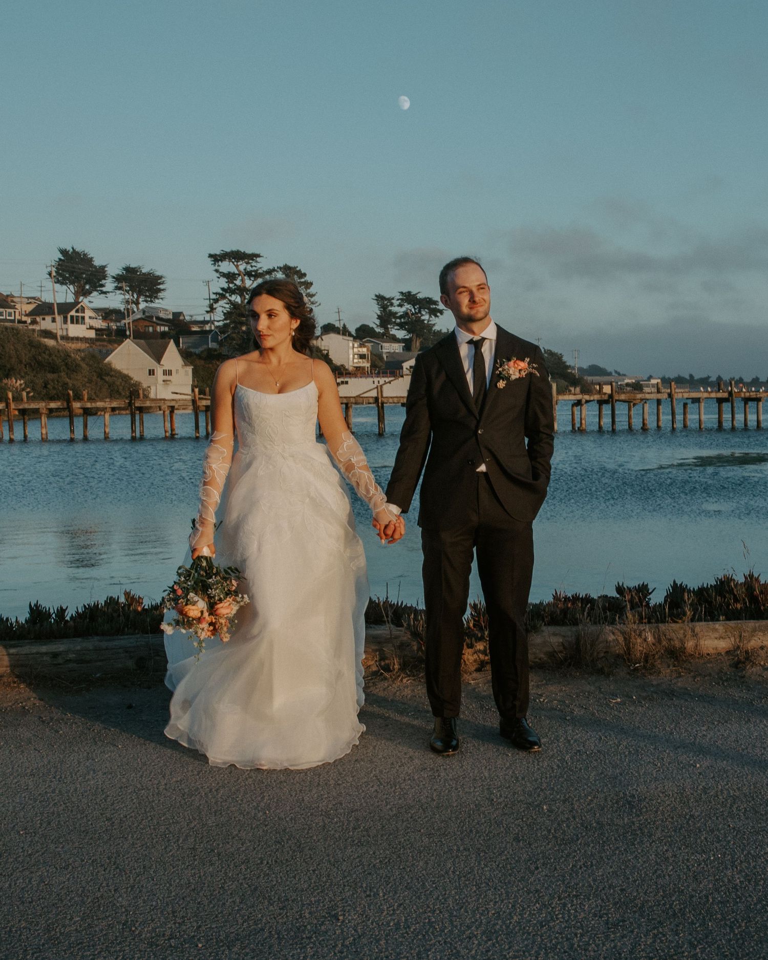 Wedding couple walking along waterfront at sunset with pier in background.