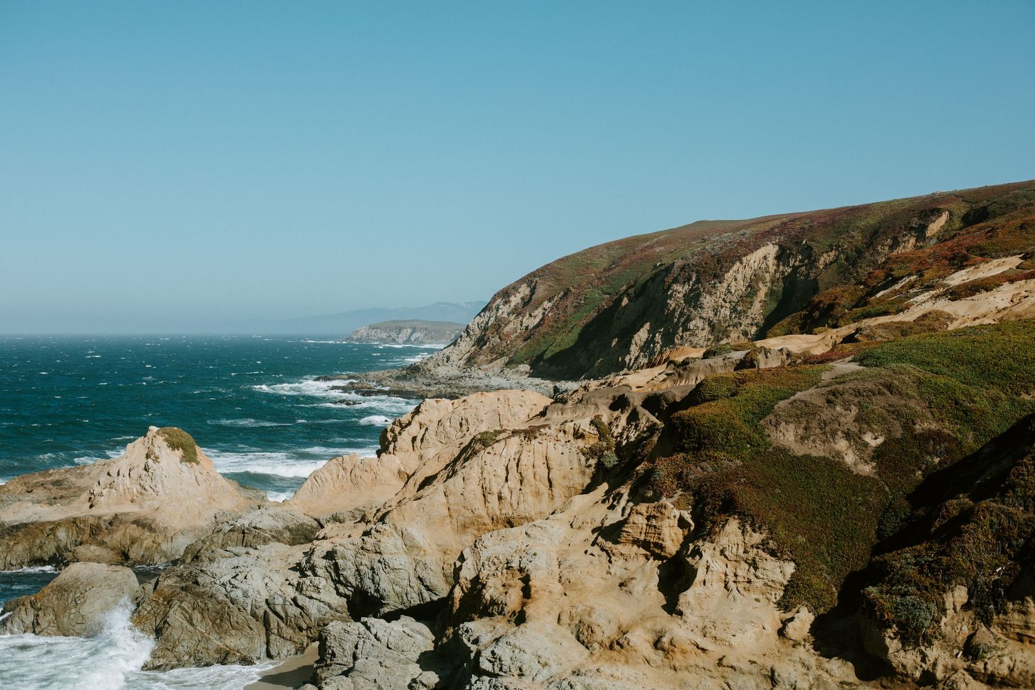 Dramatic coastal cliffs and rocky shoreline along the Pacific Ocean under clear blue skies.