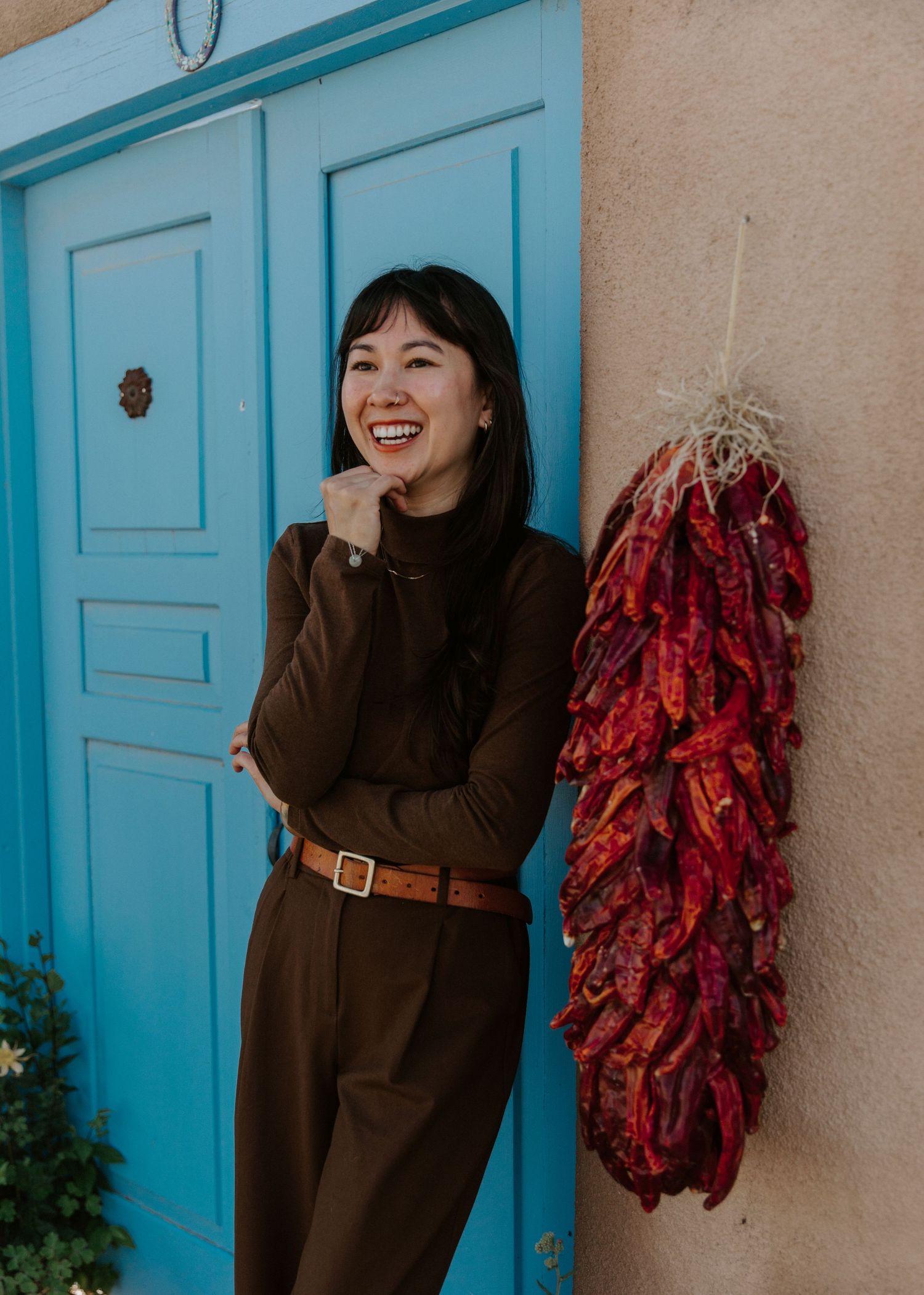 Smiling woman in brown turtleneck and trousers poses by blue door with red chile ristra in Santa Fe.