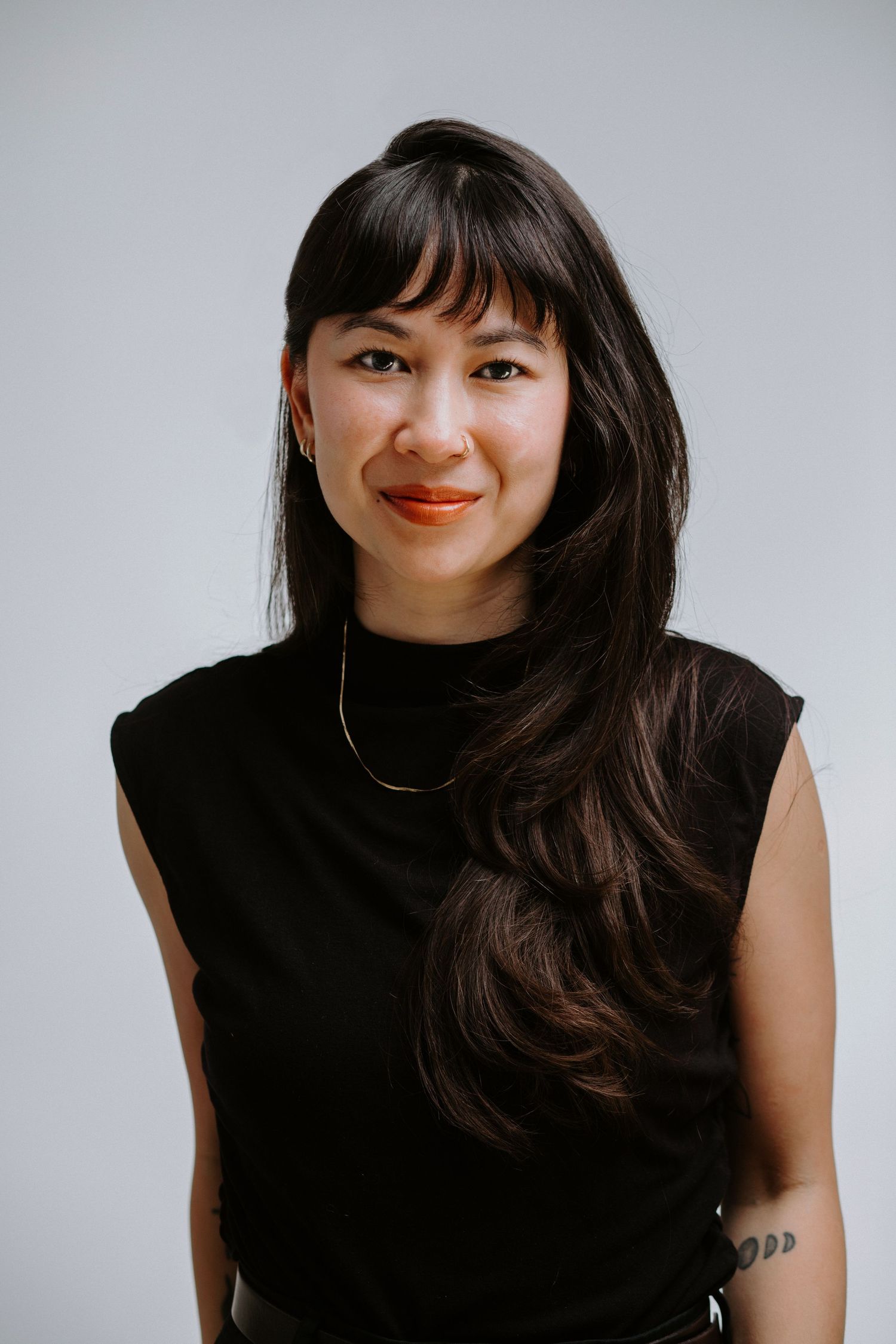 A smiling young woman with long dark hair and bangs wearing a black sleeveless top and delicate necklace against a gray background.