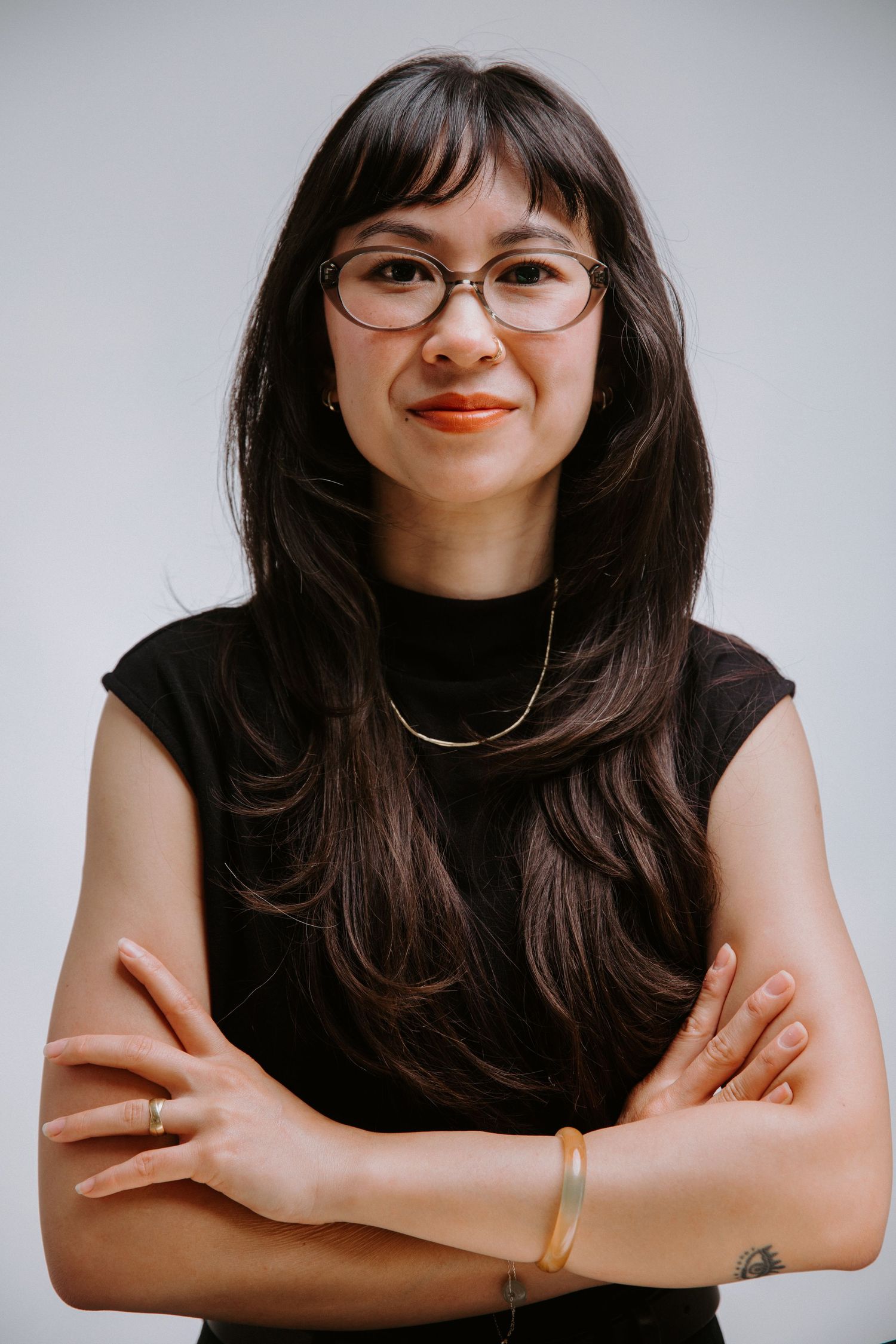 Young Asian woman with glasses and long dark hair, wearing black sleeveless top, arms crossed, smiling confidently.