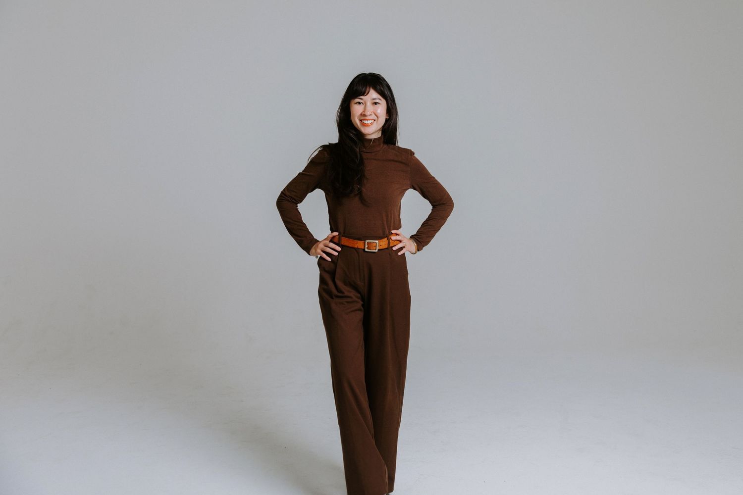 Woman in brown turtleneck and wide-leg trousers with tan belt posing confidently on white background.