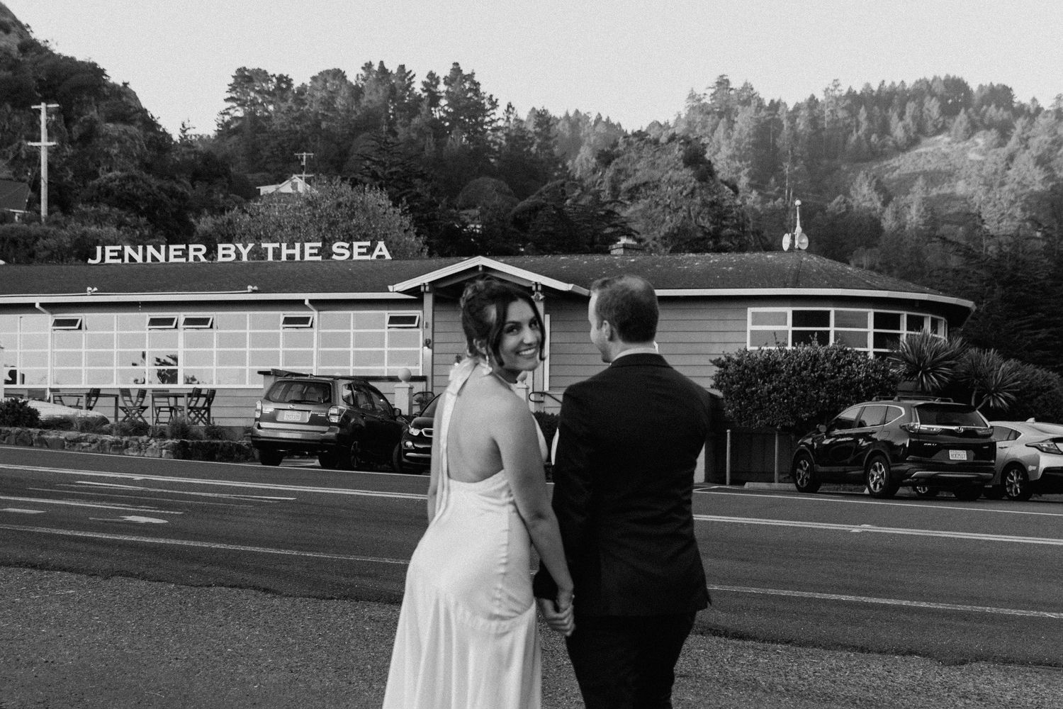 A black and white wedding photo shows a couple walking together outside a seaside venue with a church steeple visible.