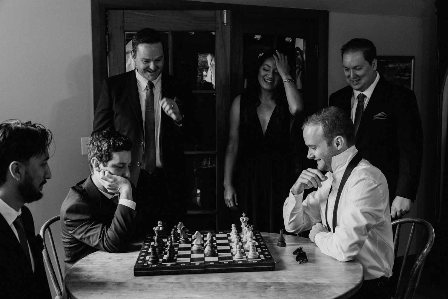 A group gathers around a chess match in a dimly lit room creating an intense atmosphere.