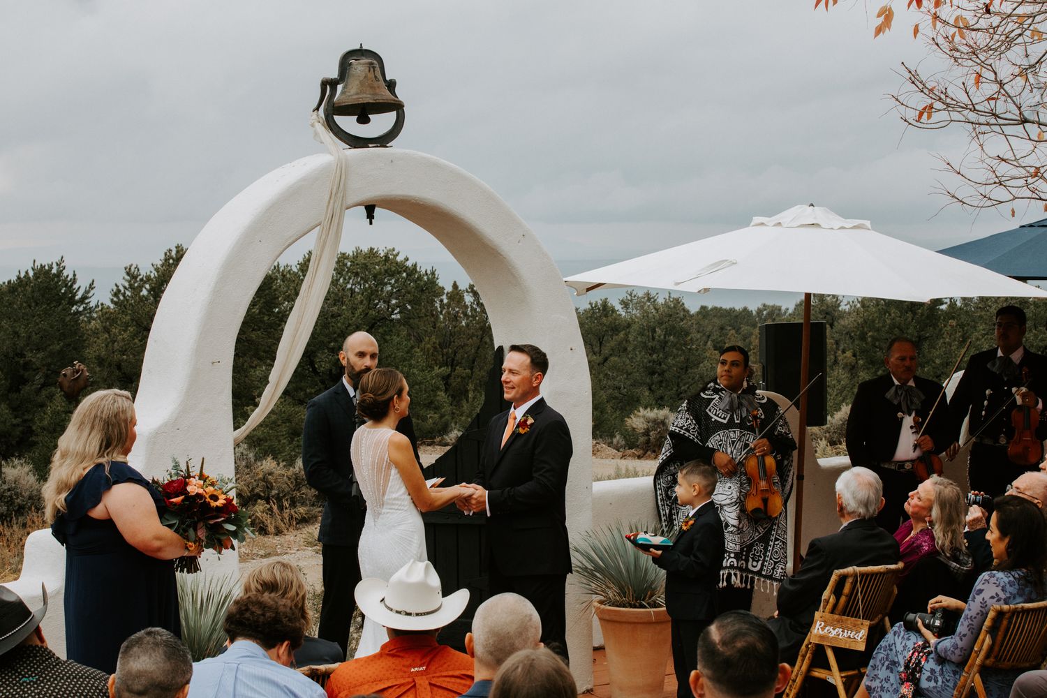 Wedding ceremony taking place under a white circular arch with guests gathered outdoors.