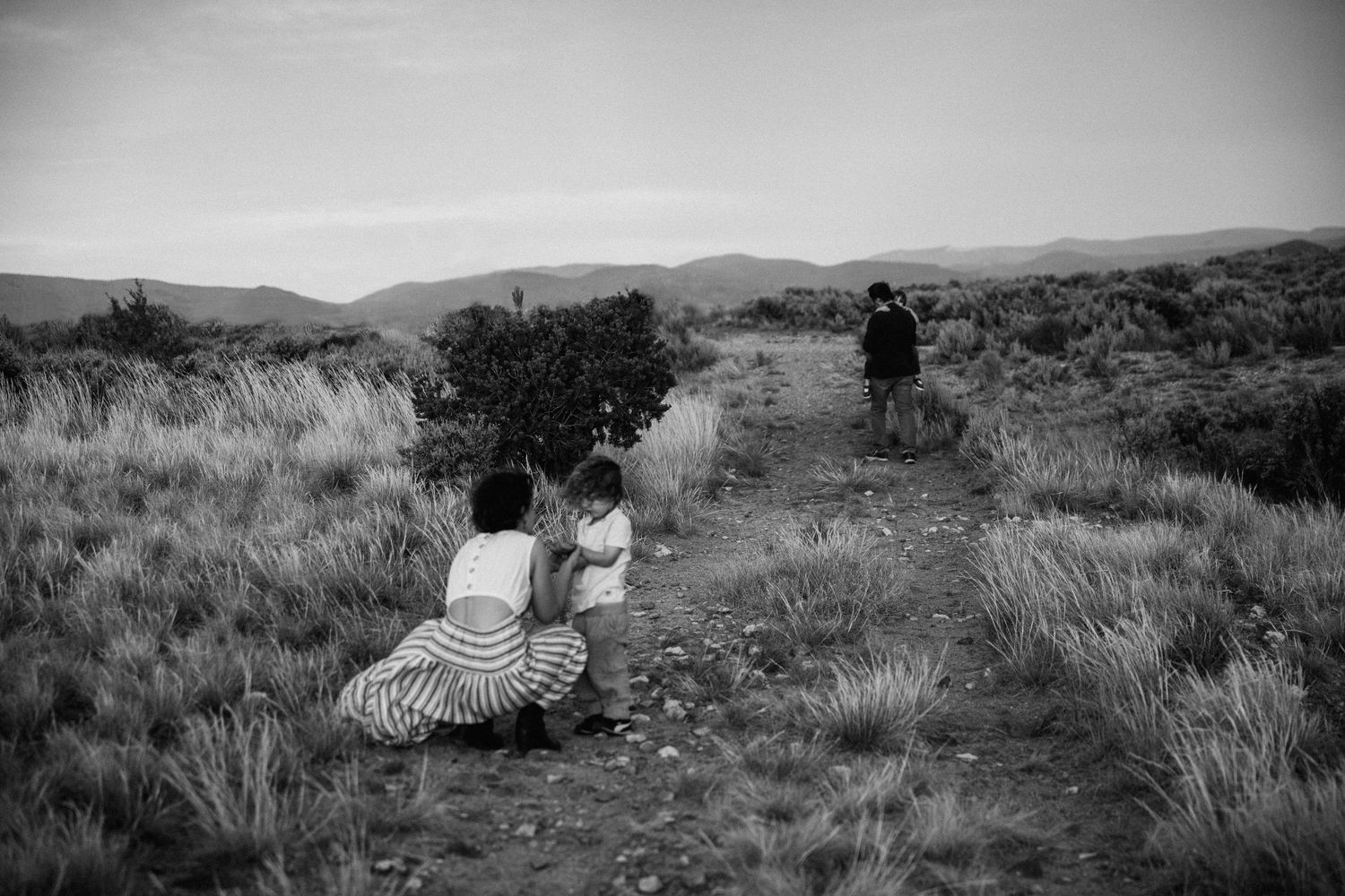 Black and white landscape photograph of people sitting in tall grass on a hillside path with mountains in the distance.