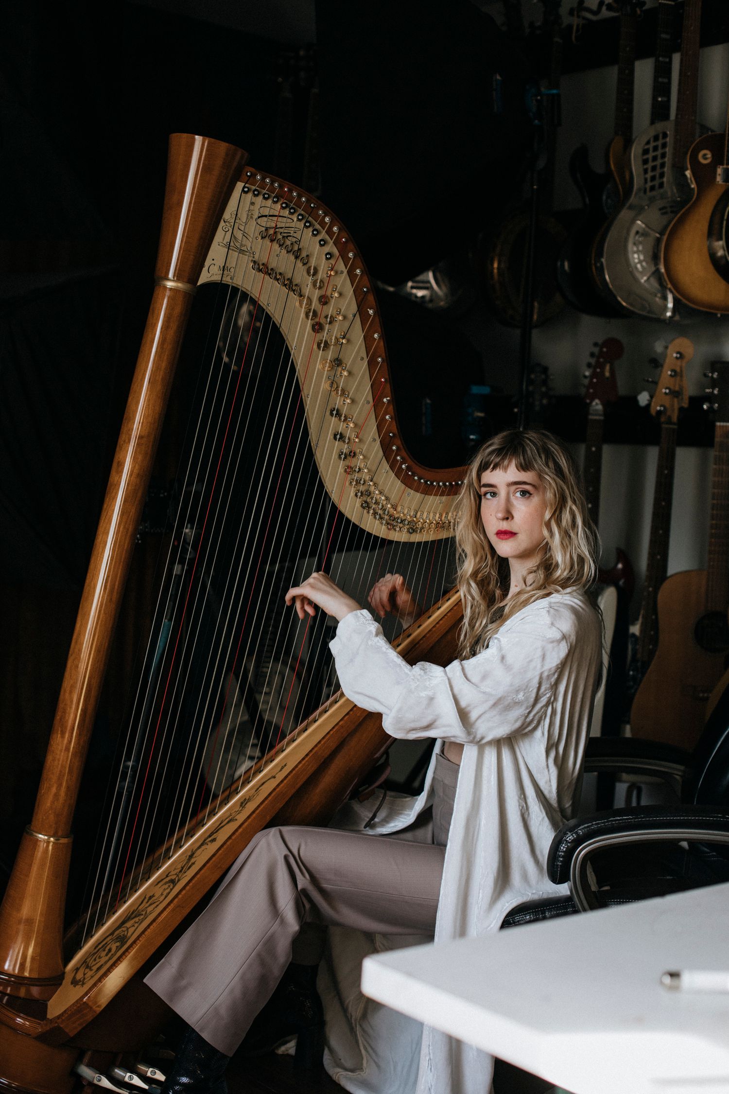 A person in a white blouse sits at an ornate wooden pedal harp in a dimly lit room.