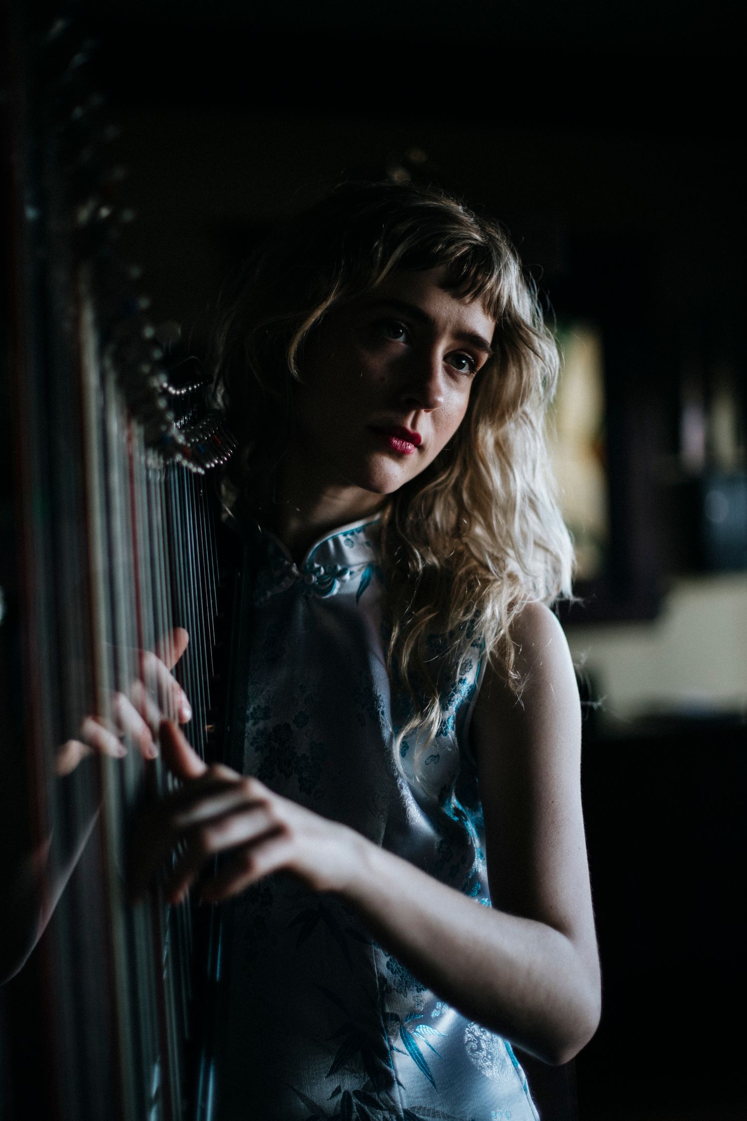 A moody portrait in dark tones shows someone in a blue dress standing by a window with dramatic lighting.