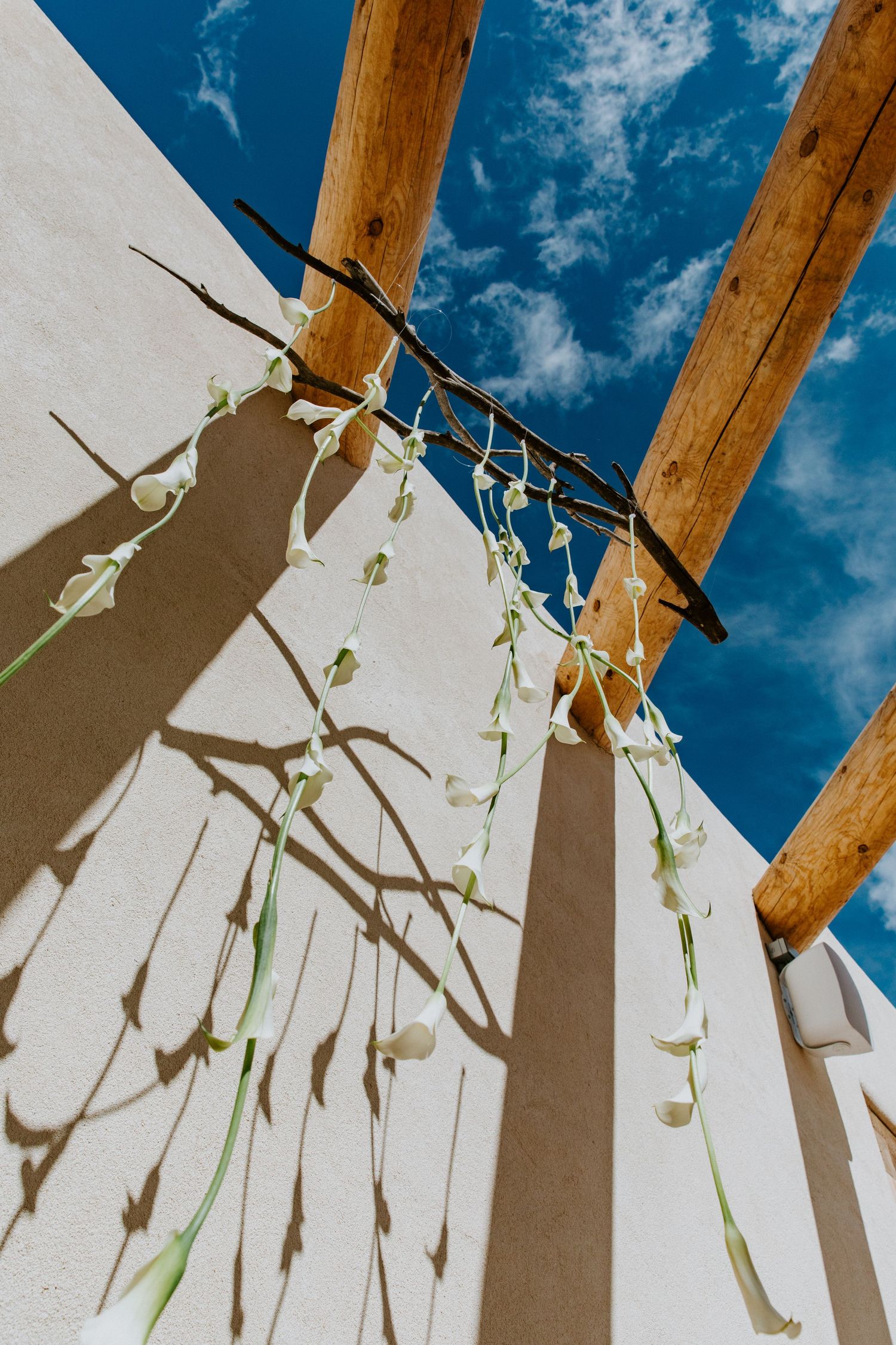 Wooden beams cast dramatic shadows against a white wall under a bright blue sky with wispy clouds.