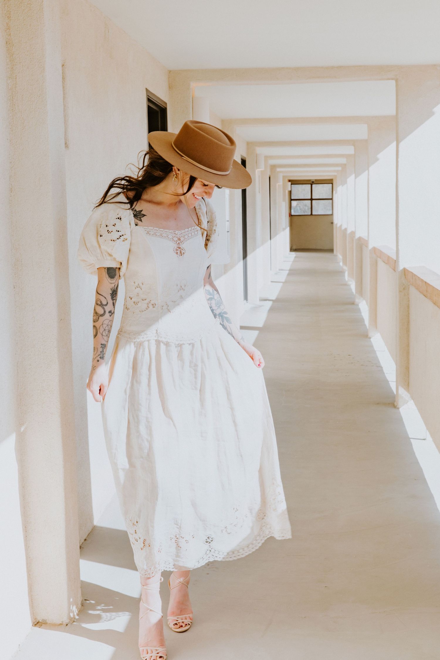 A person in a white lace dress and brown hat walks down a sunlit hallway with white columns.