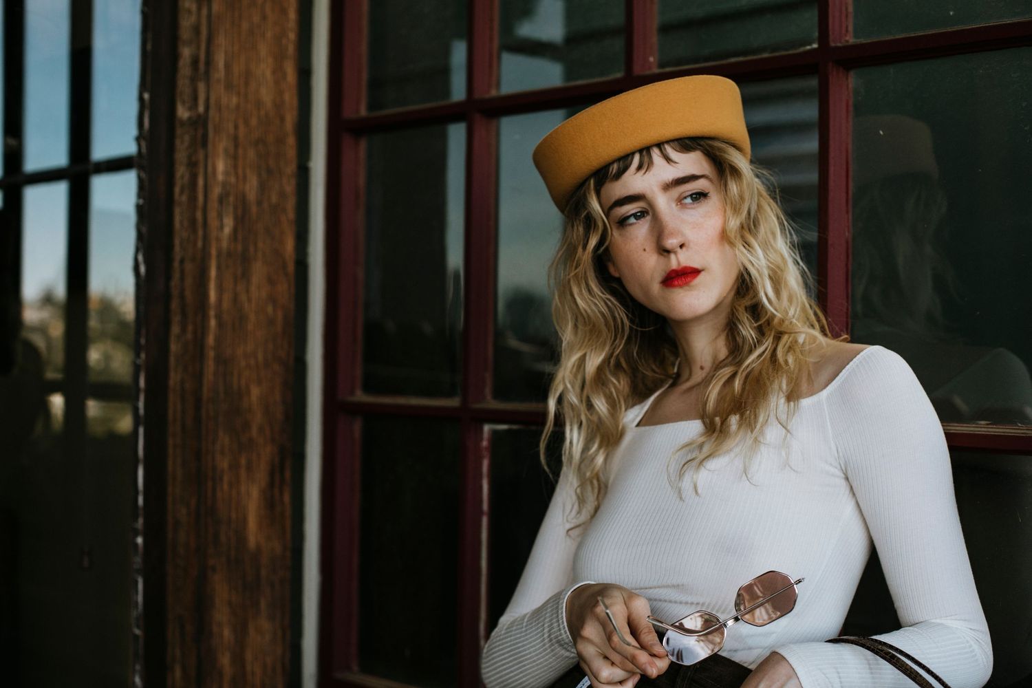 A person in a white sweater and tan beret poses against a window with warm lighting and wooden frames.