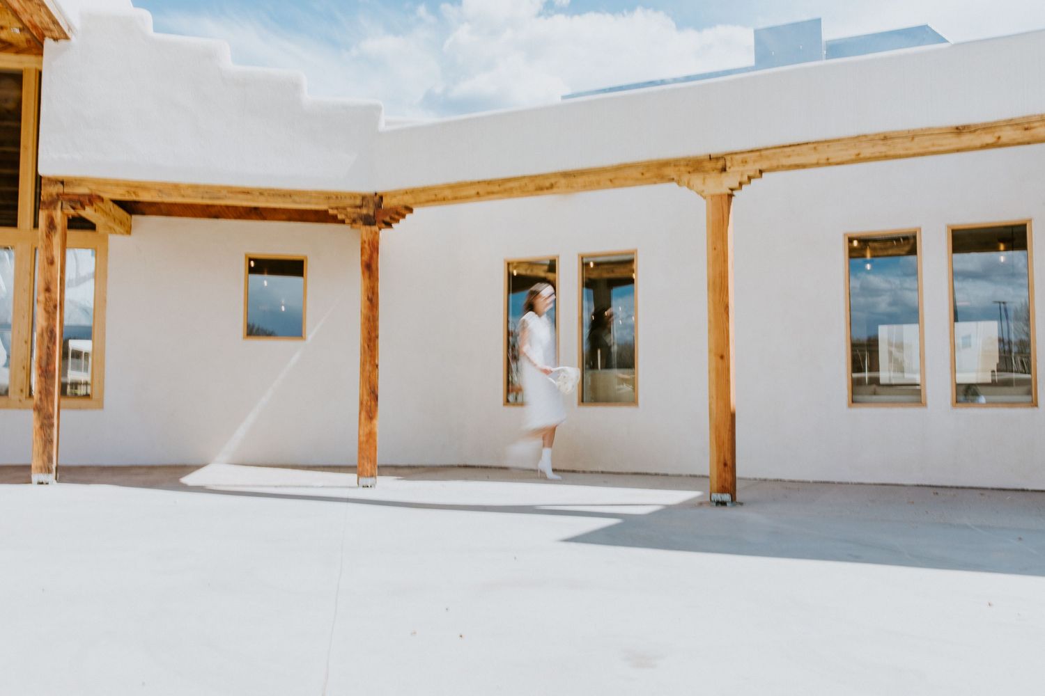 Modern covered walkway with wooden beams and white walls connects buildings in an architectural courtyard setting.