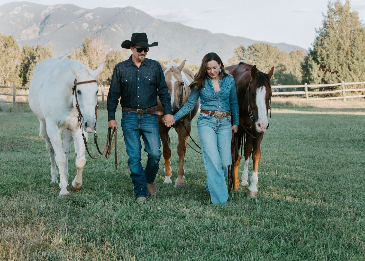 A couple walks with two horses through a grassy field with mountains in the background.