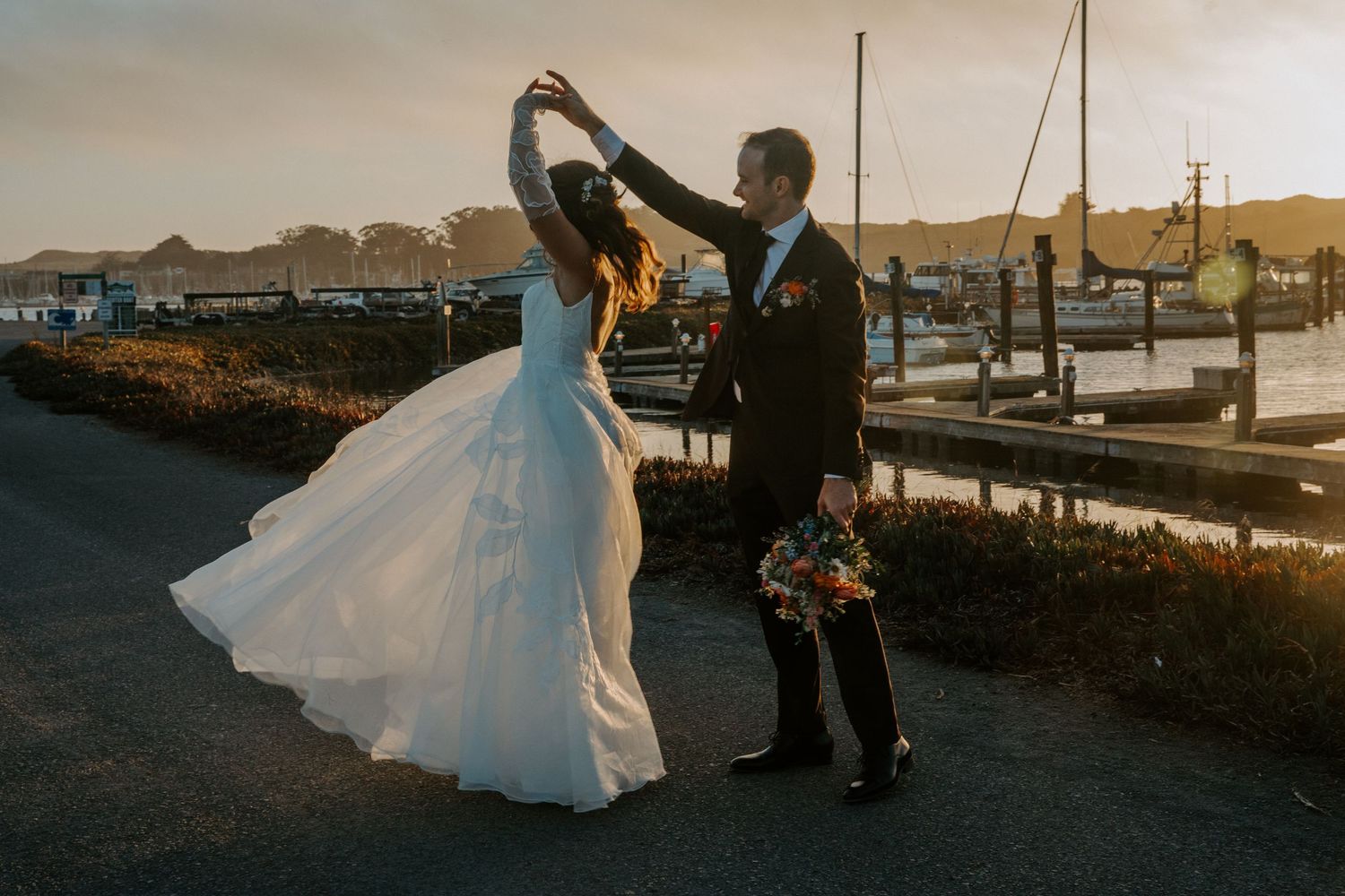 Bride and groom walking along waterfront path at sunset.