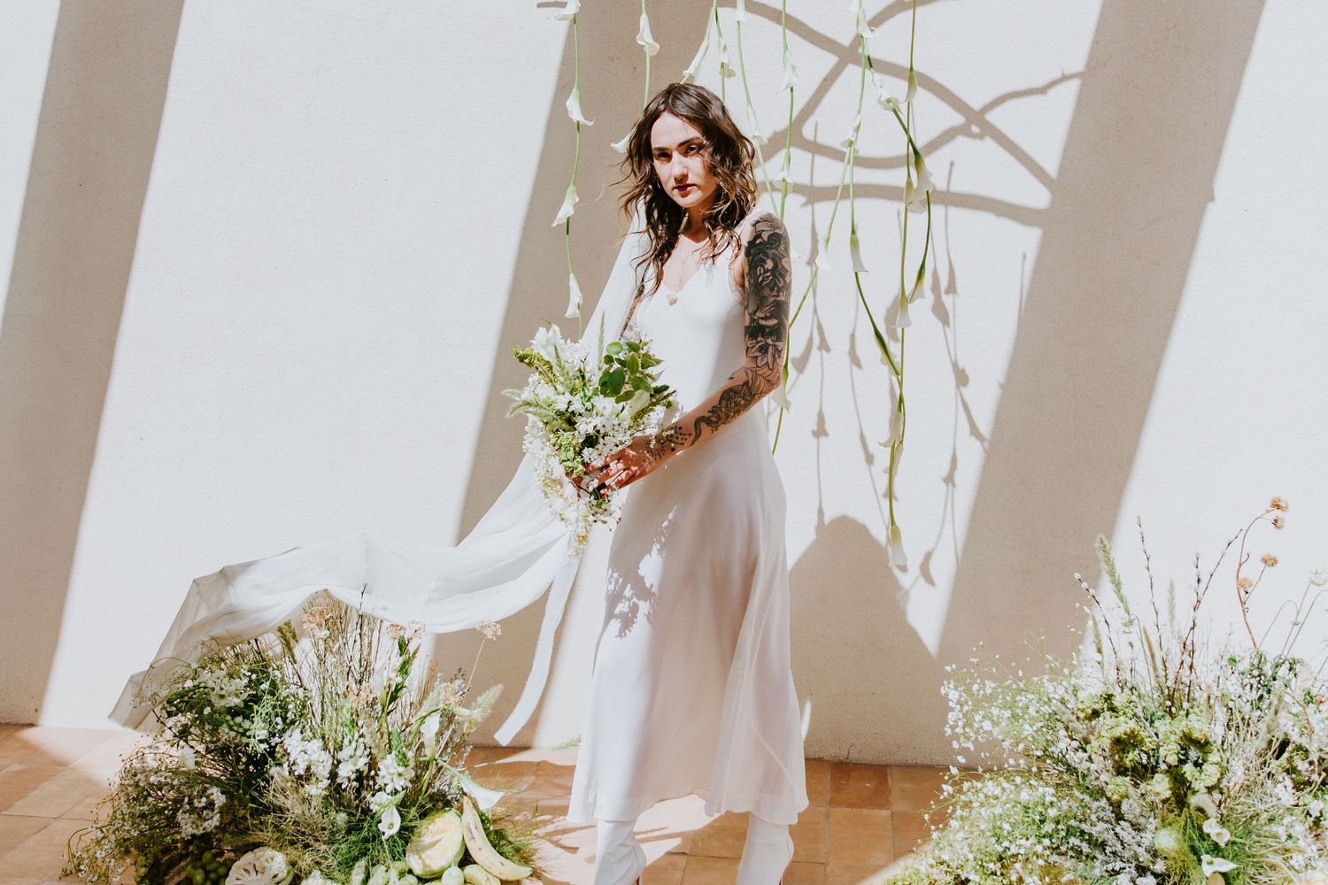 A bride in a white wedding dress holds a lush green and white wildflower bouquet against a light backdrop.