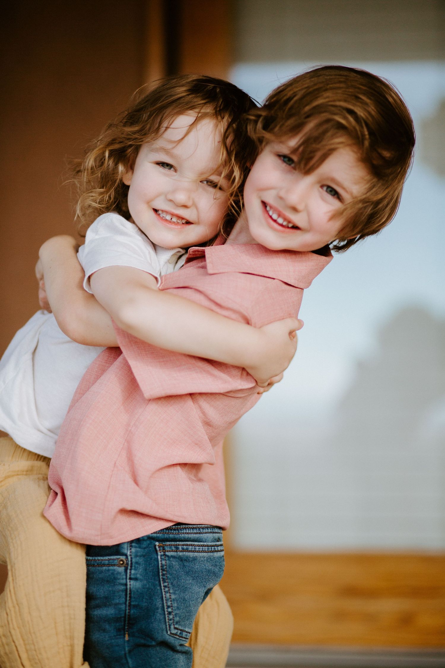 Two young children in casual clothing share a warm embrace against a light background.