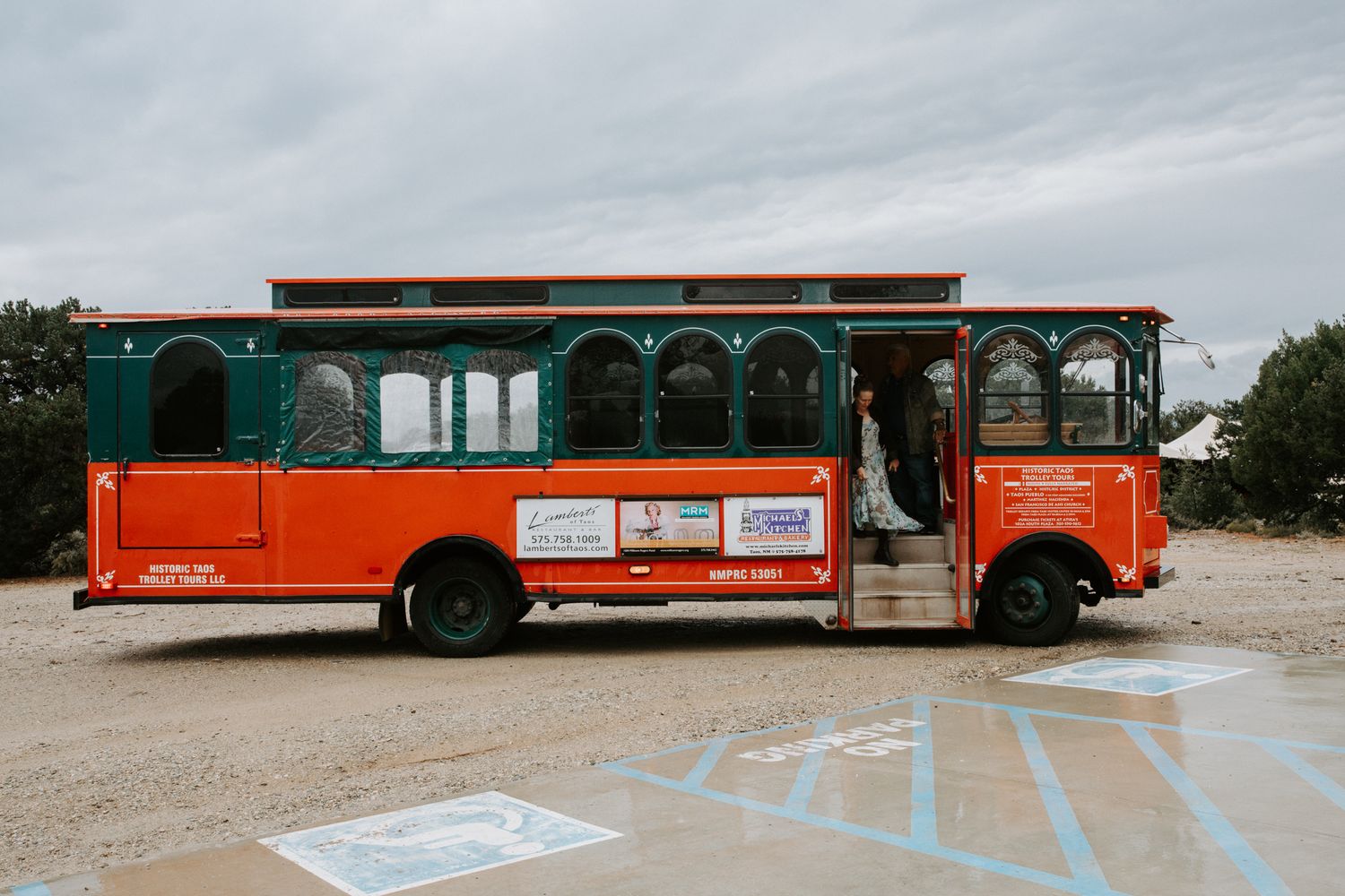 Long orange and teal colored buses parked in a line on a dirt lot.