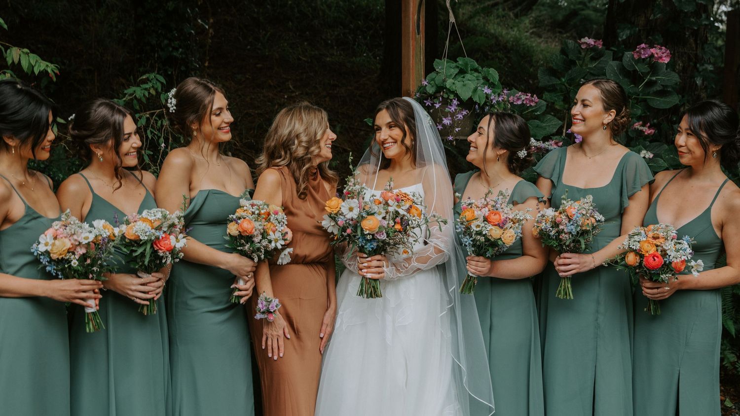 A wedding party stands together holding colorful bouquets in sage green dresses against a lush outdoor backdrop.