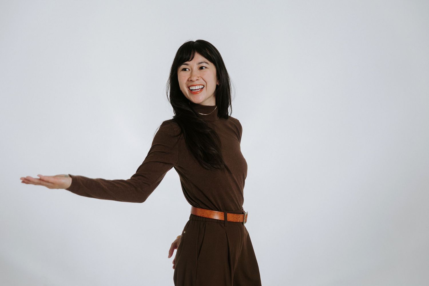 Smiling Asian woman in brown turtleneck and skirt gesturing with open hand on white background.