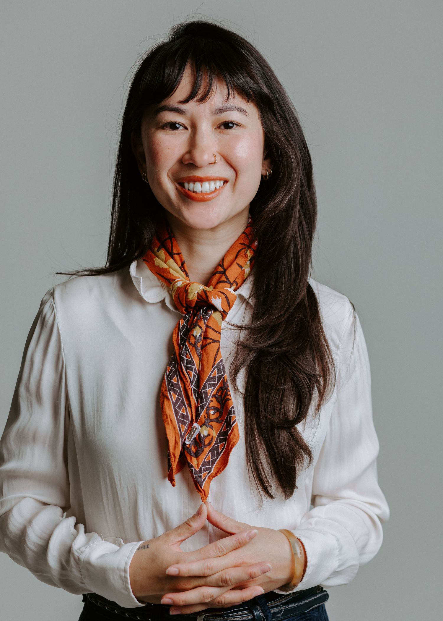 Smiling young woman with long dark hair wearing a white blouse and orange patterned scarf, posing against a gray background.