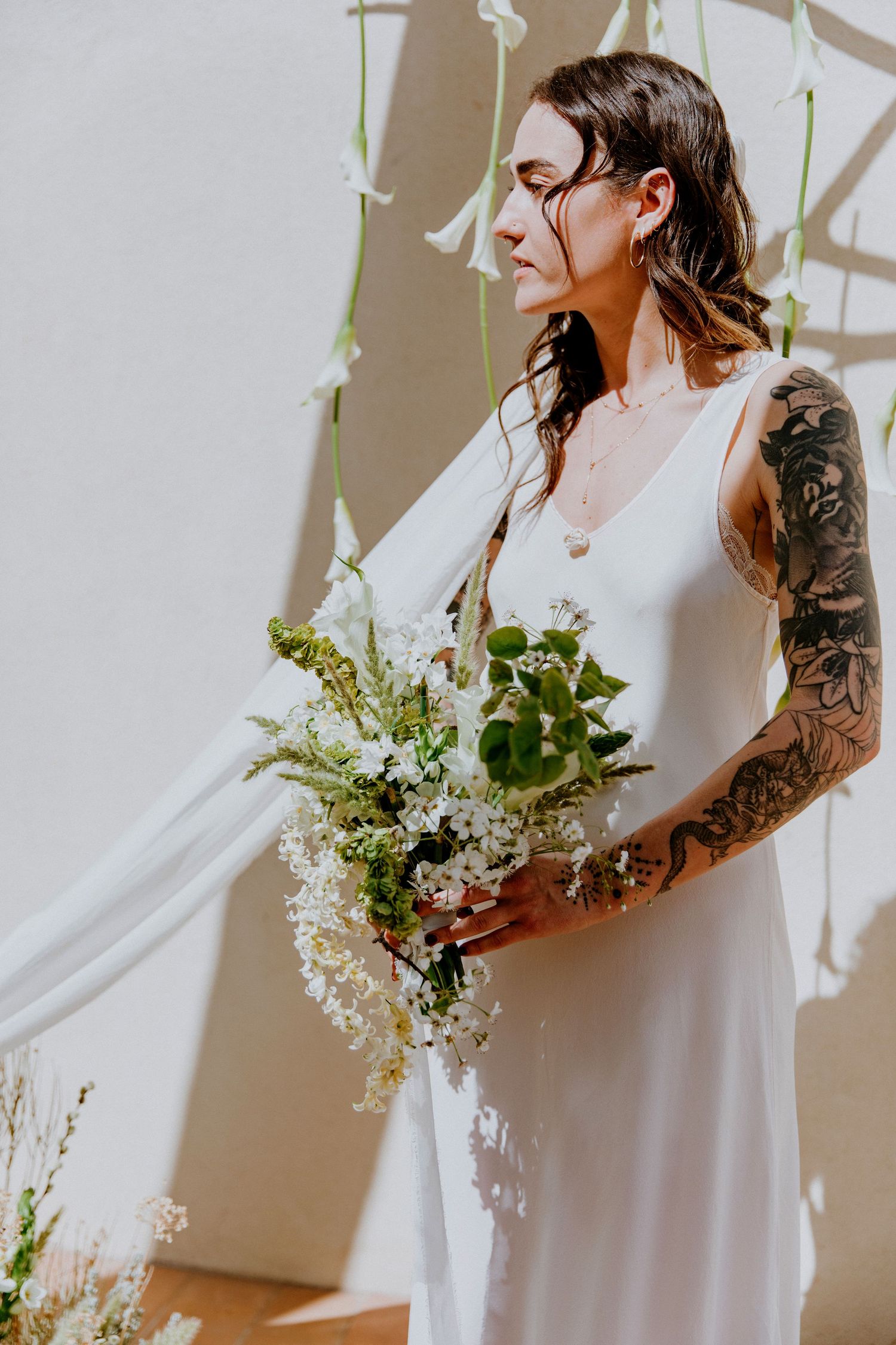 Bride in flowing white dress holds delicate bouquet of white flowers against neutral wall in elegant bridal portrait series.