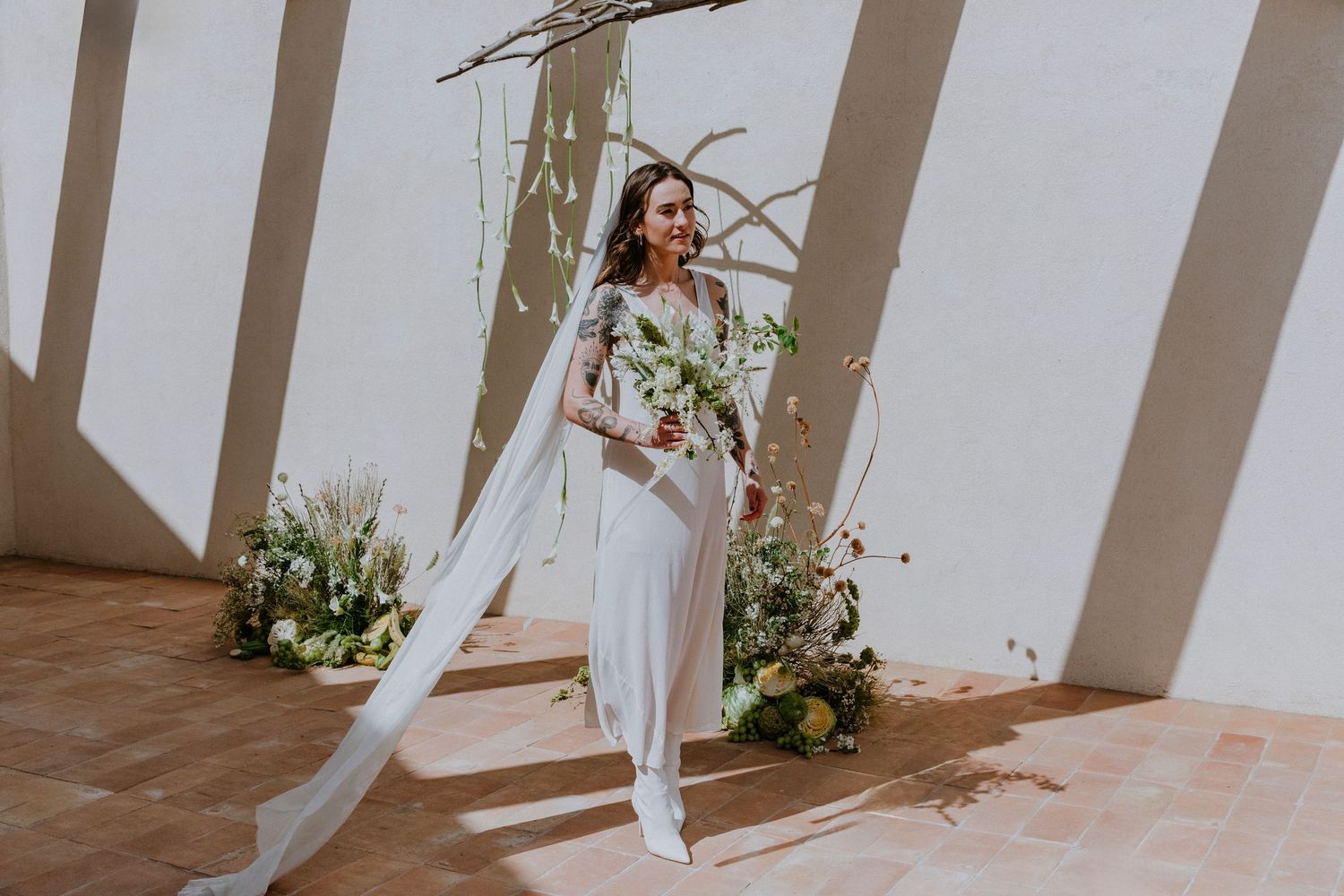 Modern bridal portrait in white jumpsuit standing against white wall with floral arrangements and dramatic shadows.