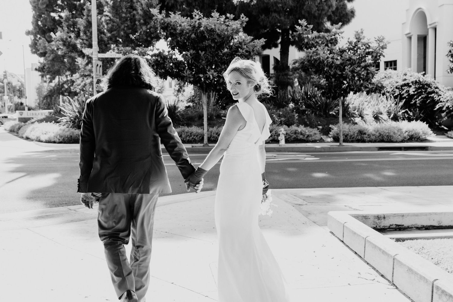 Black and white photo of a bride in white gown holding hands with groom in suit, walking outdoors.
