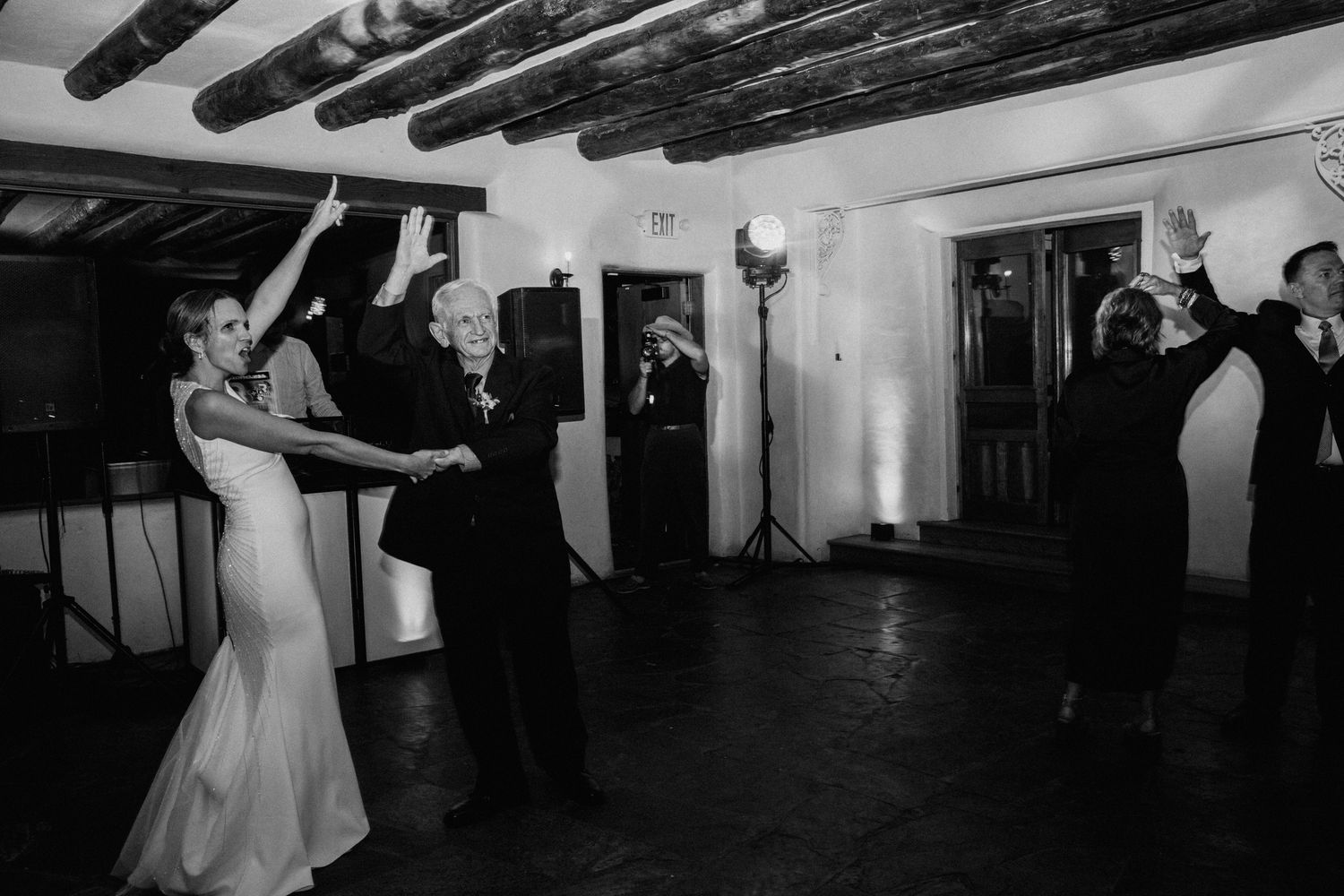 Black and white photo of couples dancing together at a wedding reception in a rustic venue with exposed beams.
