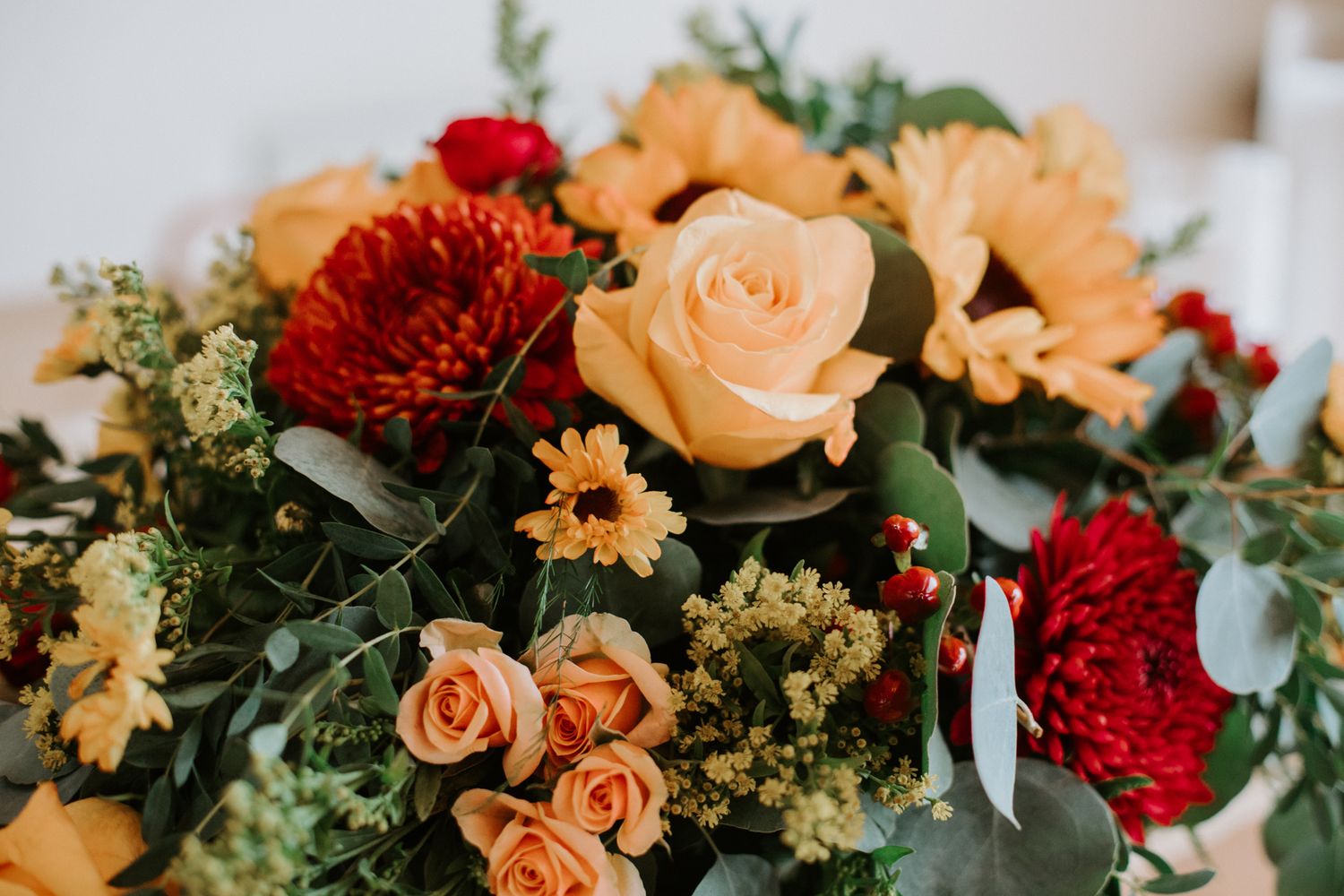 A floral arrangement featuring peach roses, red carnations, and greenery in warm autumn colors.