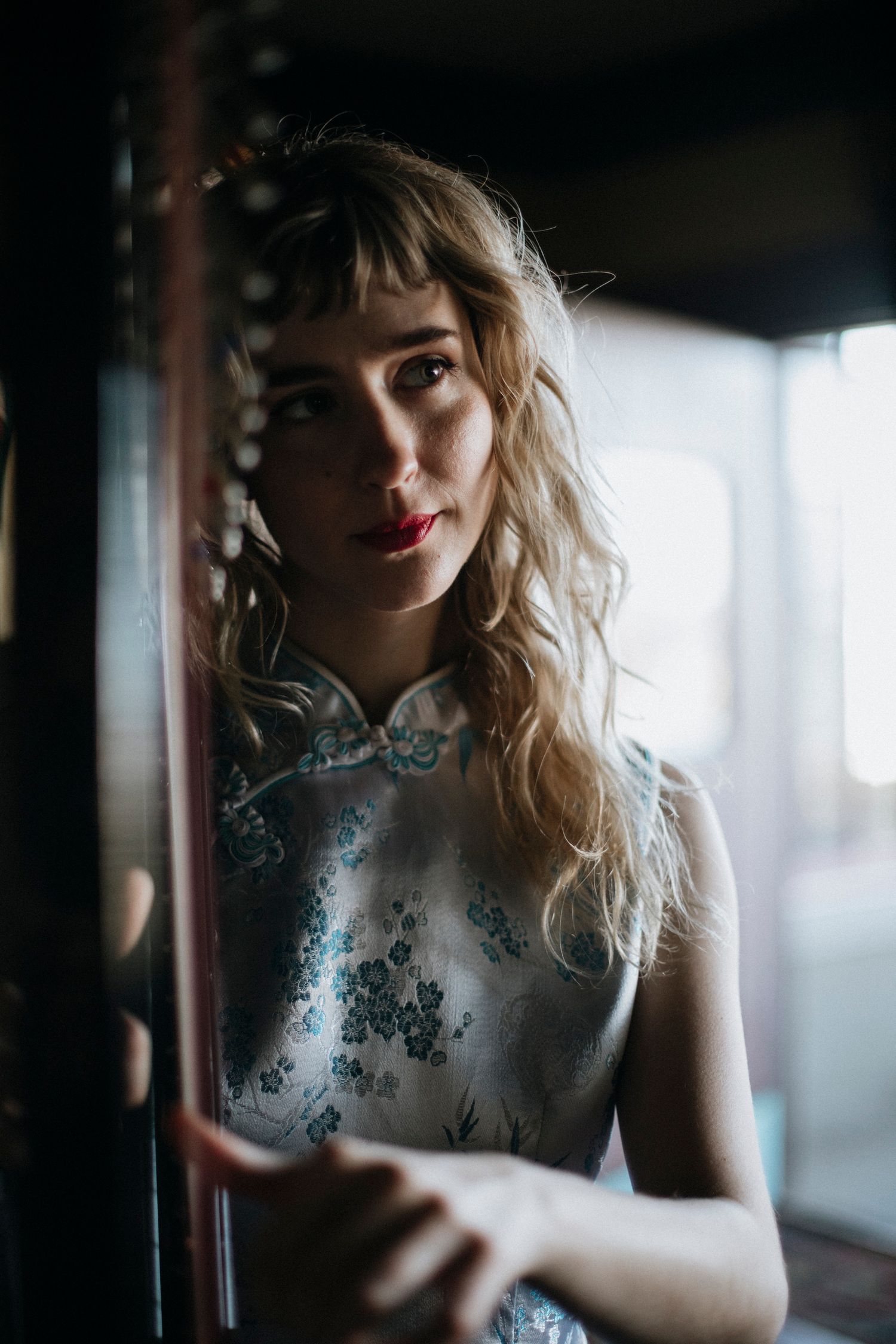 A moody portrait of someone in a light blue floral cheongsam dress standing near a window in soft natural light.