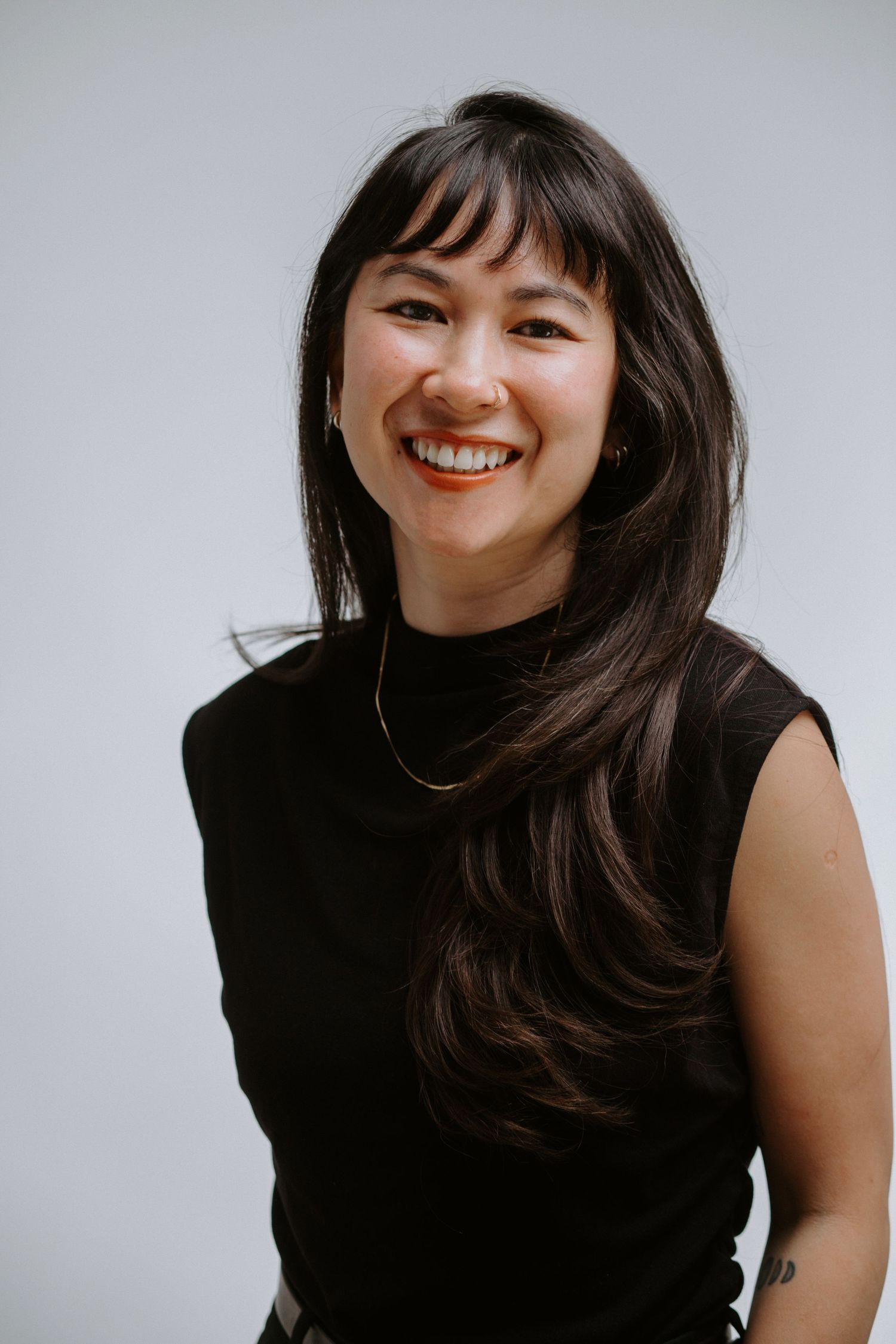 Happy Asian woman with long dark hair and bangs, wearing black sleeveless turtleneck, bright smile.