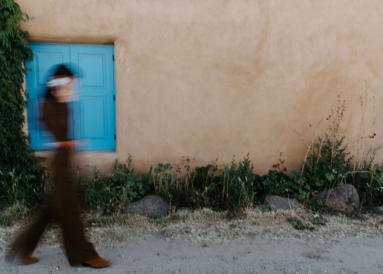 Blurred figure walking past a rustic adobe wall with a turquoise window surrounded by wild greenery.