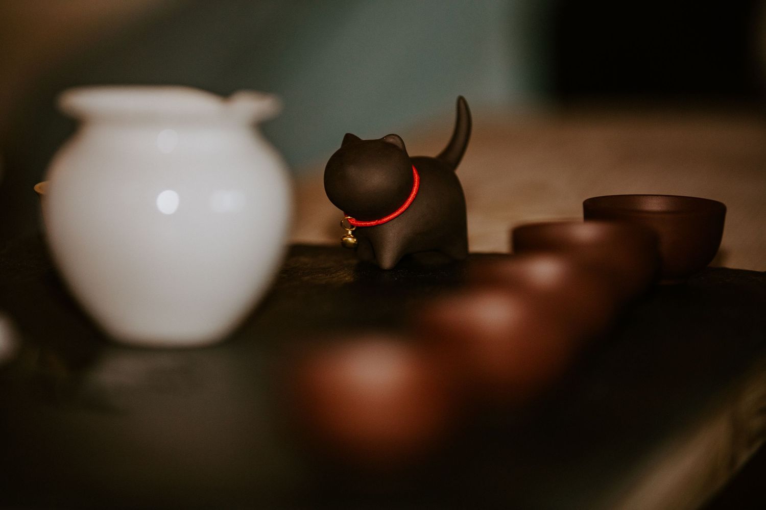 A small ceramic black cat figurine with red collar sits among brown clay tea cups on a dark wooden surface.