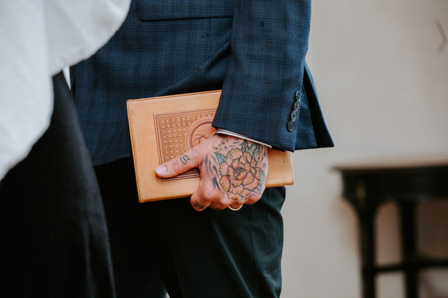 Close-up shot of someone in a navy suit holding a tan leather wallet and glass tumbler with whiskey.