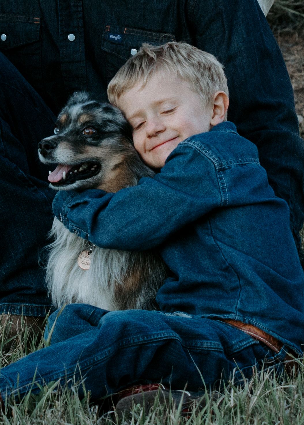 A young child in denim hugs a happy Australian Shepherd dog while sitting in grass.