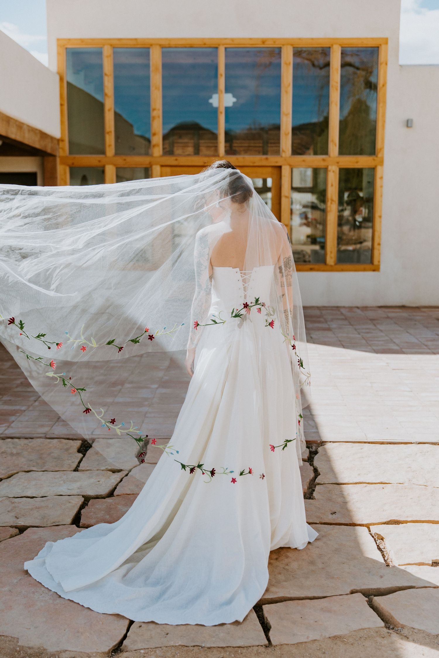 A flowing white wedding dress with a long veil blowing in the wind against a stone patio and building exterior.
