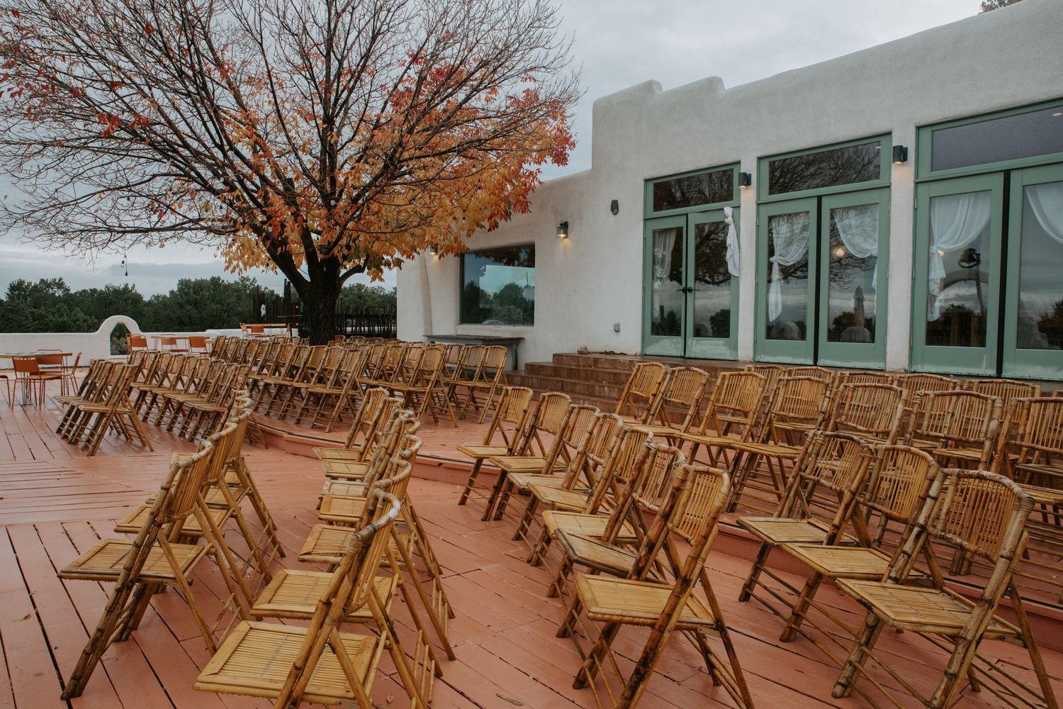 Empty wooden folding chairs arranged in rows on a terrace with autumn trees in the background.