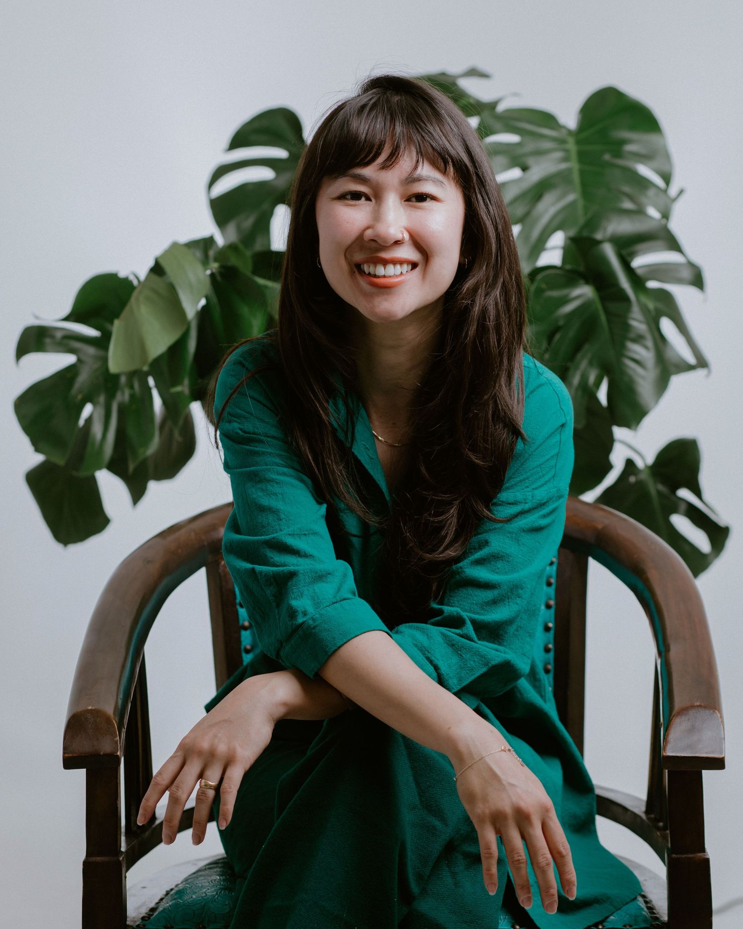 Smiling woman in teal dress seated in wooden chair with monstera plant backdrop.