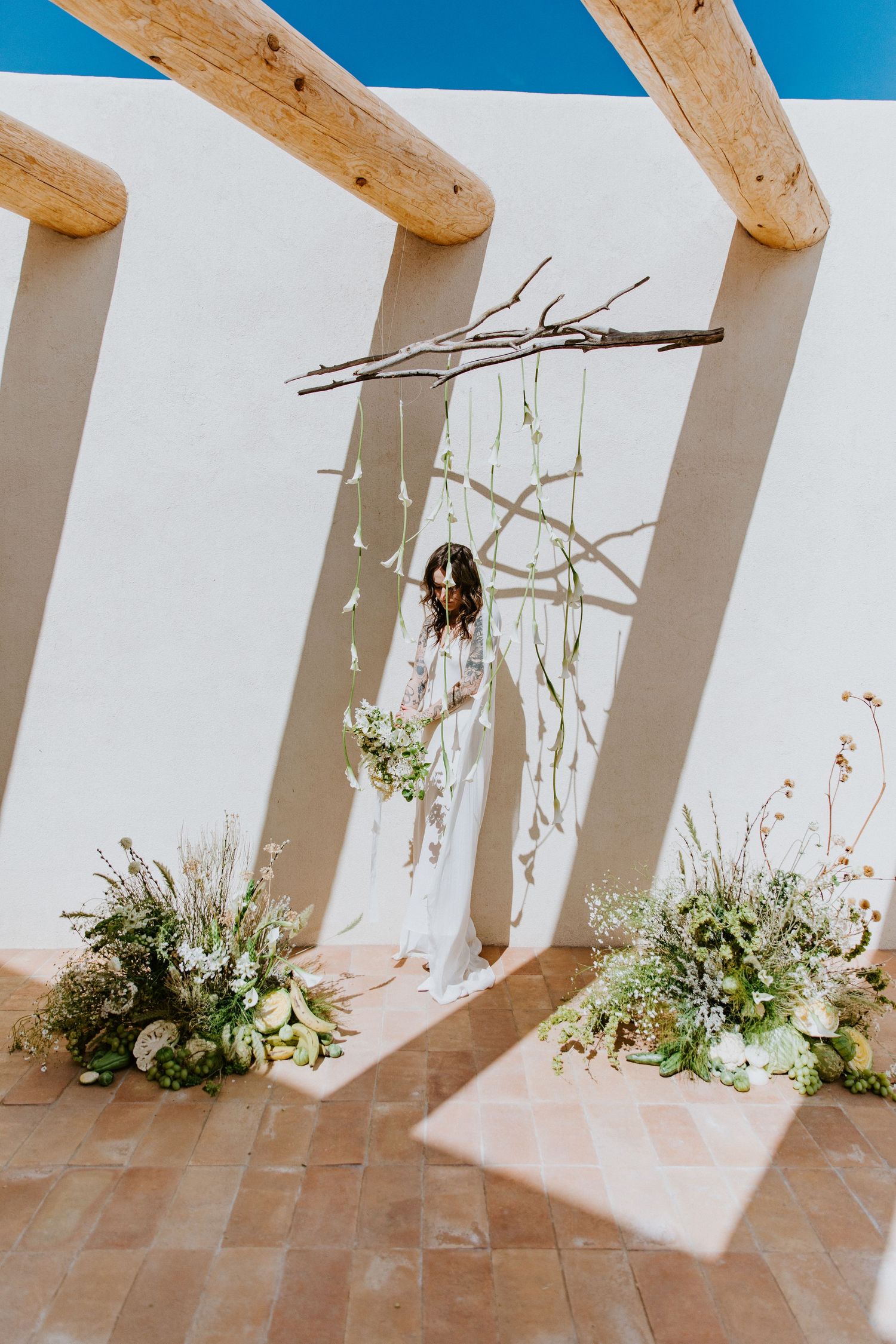 Terra cotta tiled floor meets white walls with wooden beams casting shadows and small plants growing along the base.