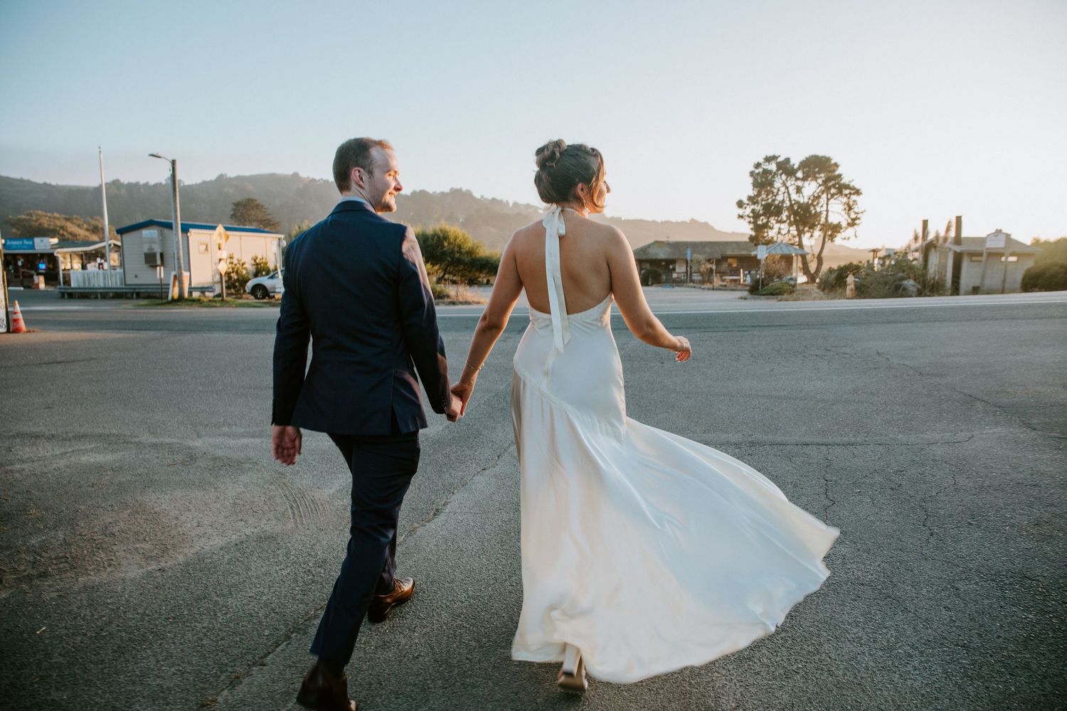 A couple in wedding attire walks together at sunset, with the bride's white dress flowing in the breeze.