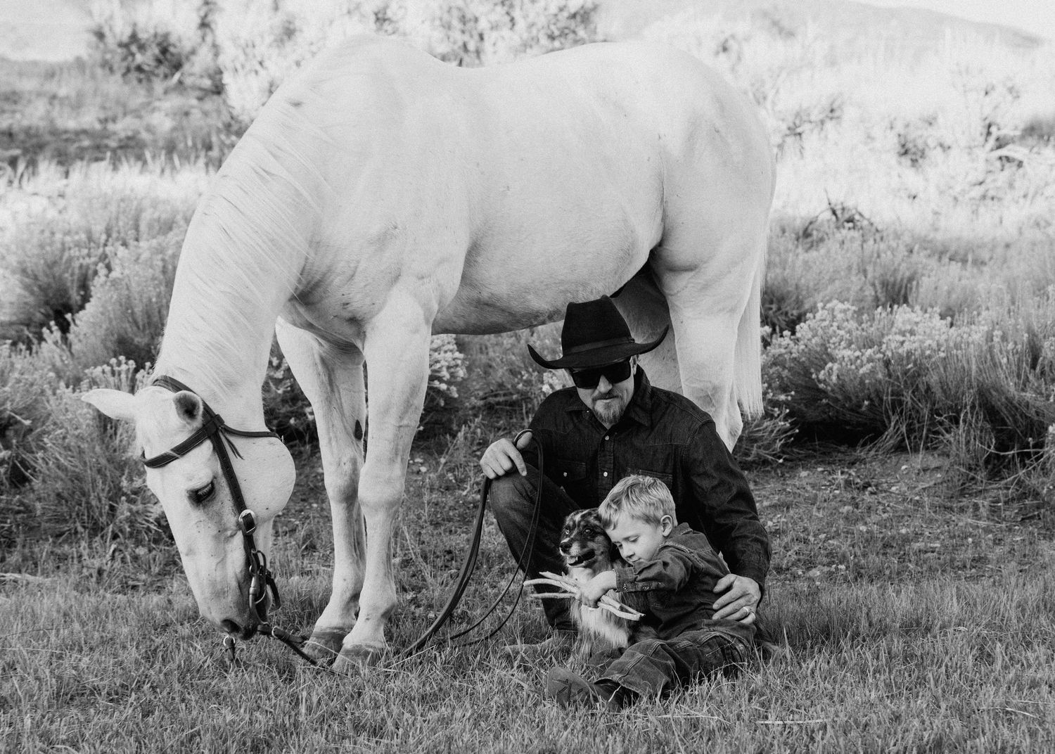 A group relaxing in a grassy field with a white horse on a sunny day, captured in black and white.