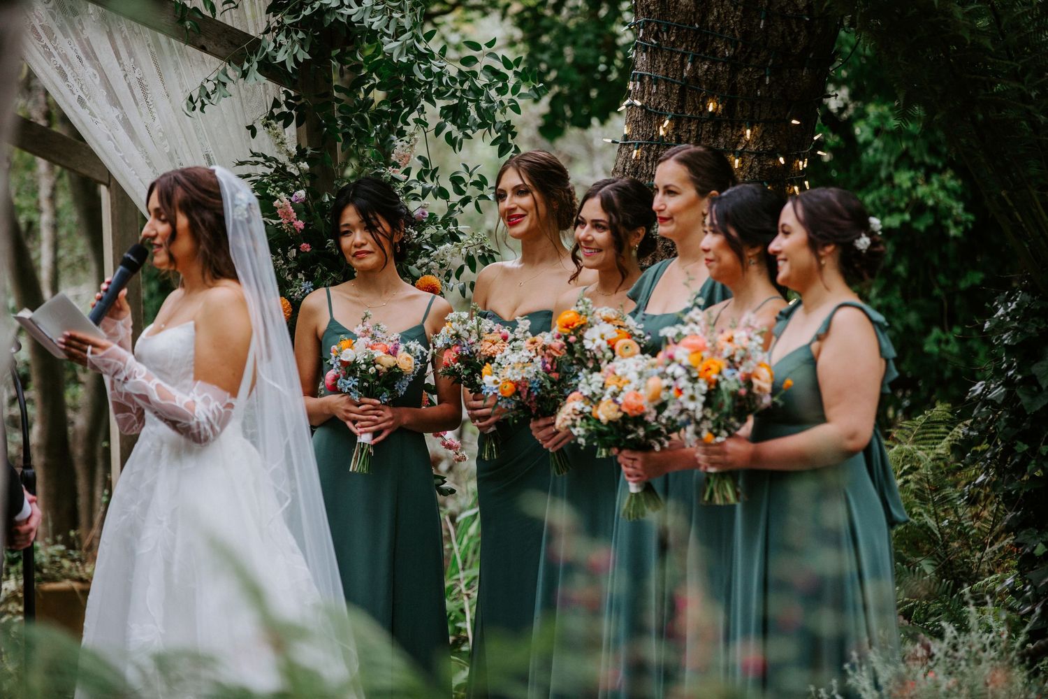A wedding party stands in a lush garden setting holding vibrant orange and pink floral bouquets.
